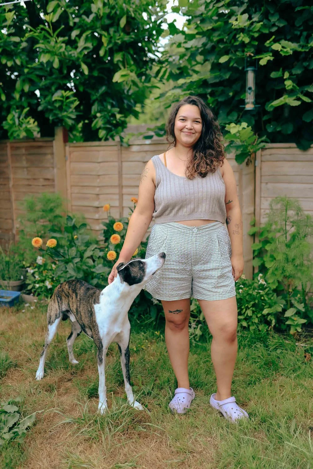 Young woman smiling in a garden with a dog, wooden fence, flowers, and lush green trees in the background.