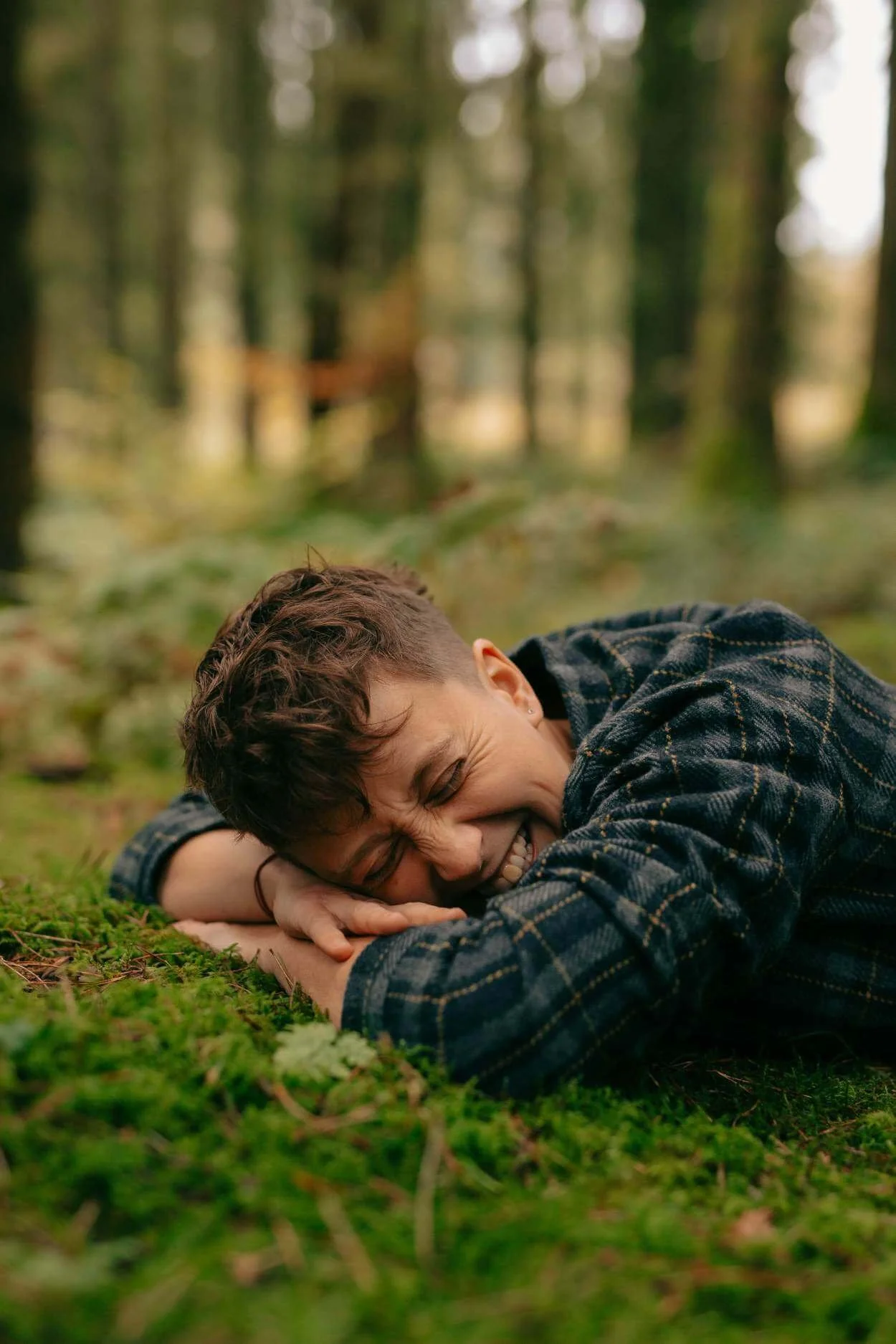 A person lying on the ground in a forest, smiling and hugging their arm, with trees blurred in the background.