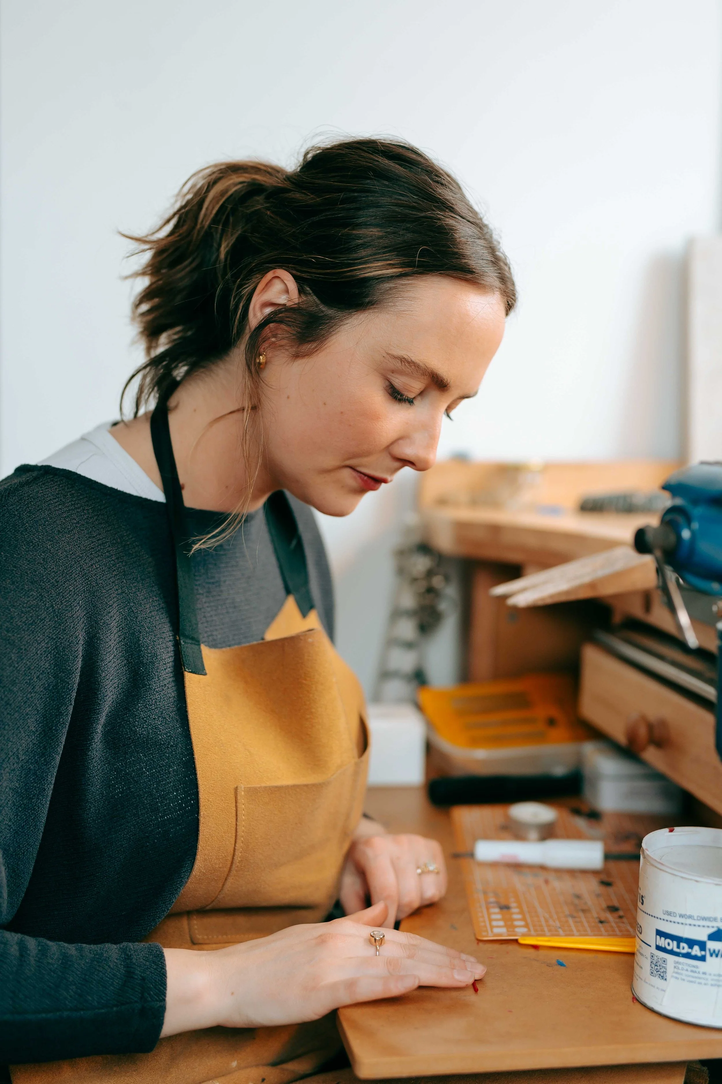 A woman wearing a black long sleeve shirt and a brown apron is working on a craft project at a wooden workbench, focusing intently on her hands and the materials in front of her.
