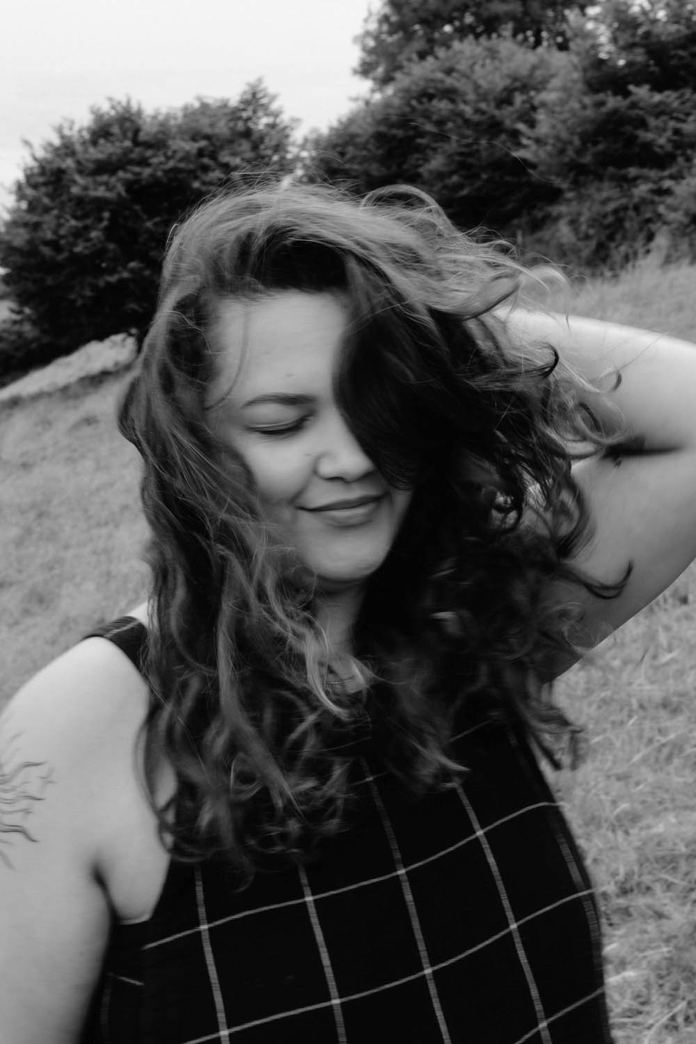 A woman with long, curly hair smiling with her eyes closed outdoors, with trees and grass in the background.