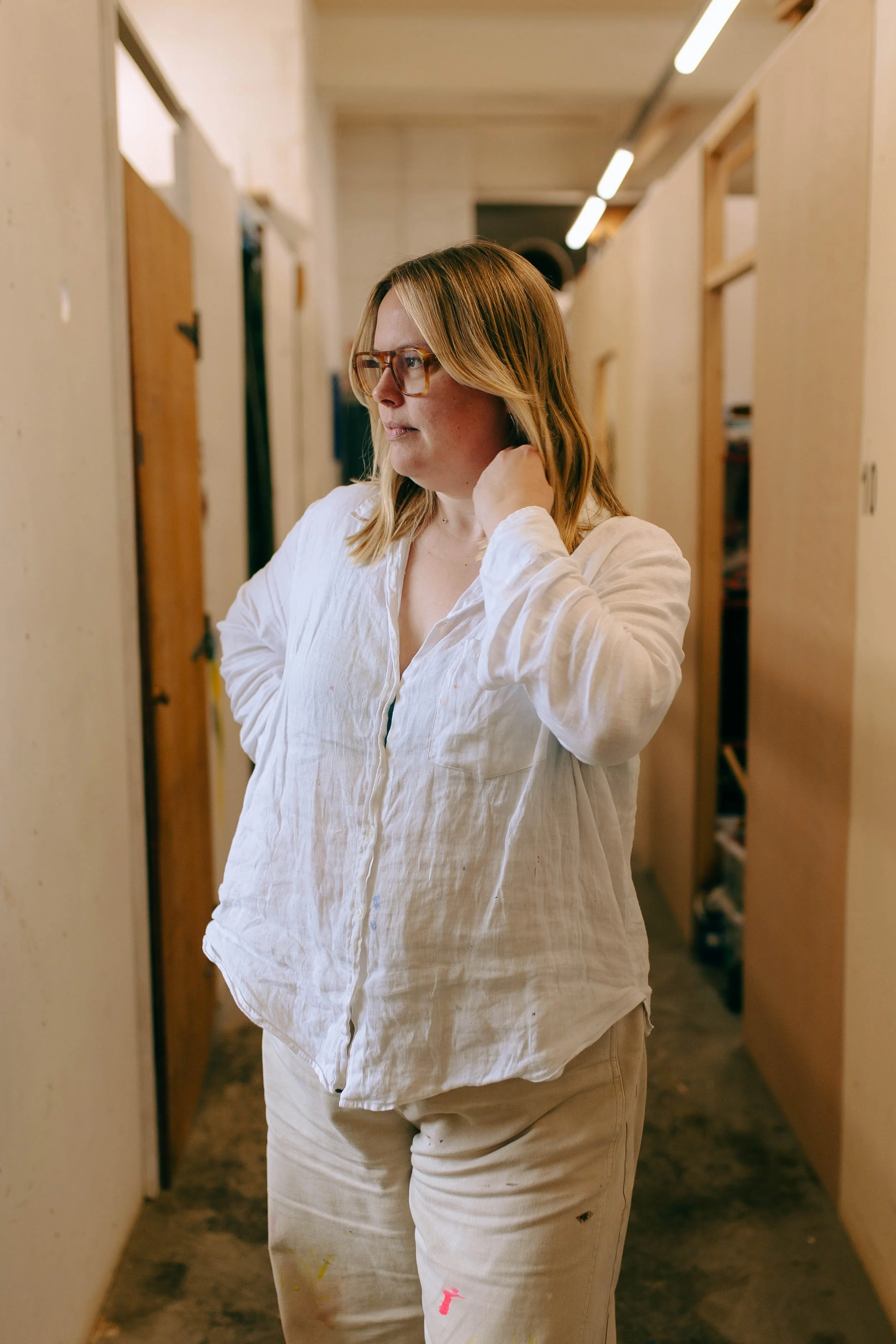 A woman with glasses and long blonde hair wearing a white shirt, standing in a hallway with wooden walls and a brown carpet, looking to the side.
