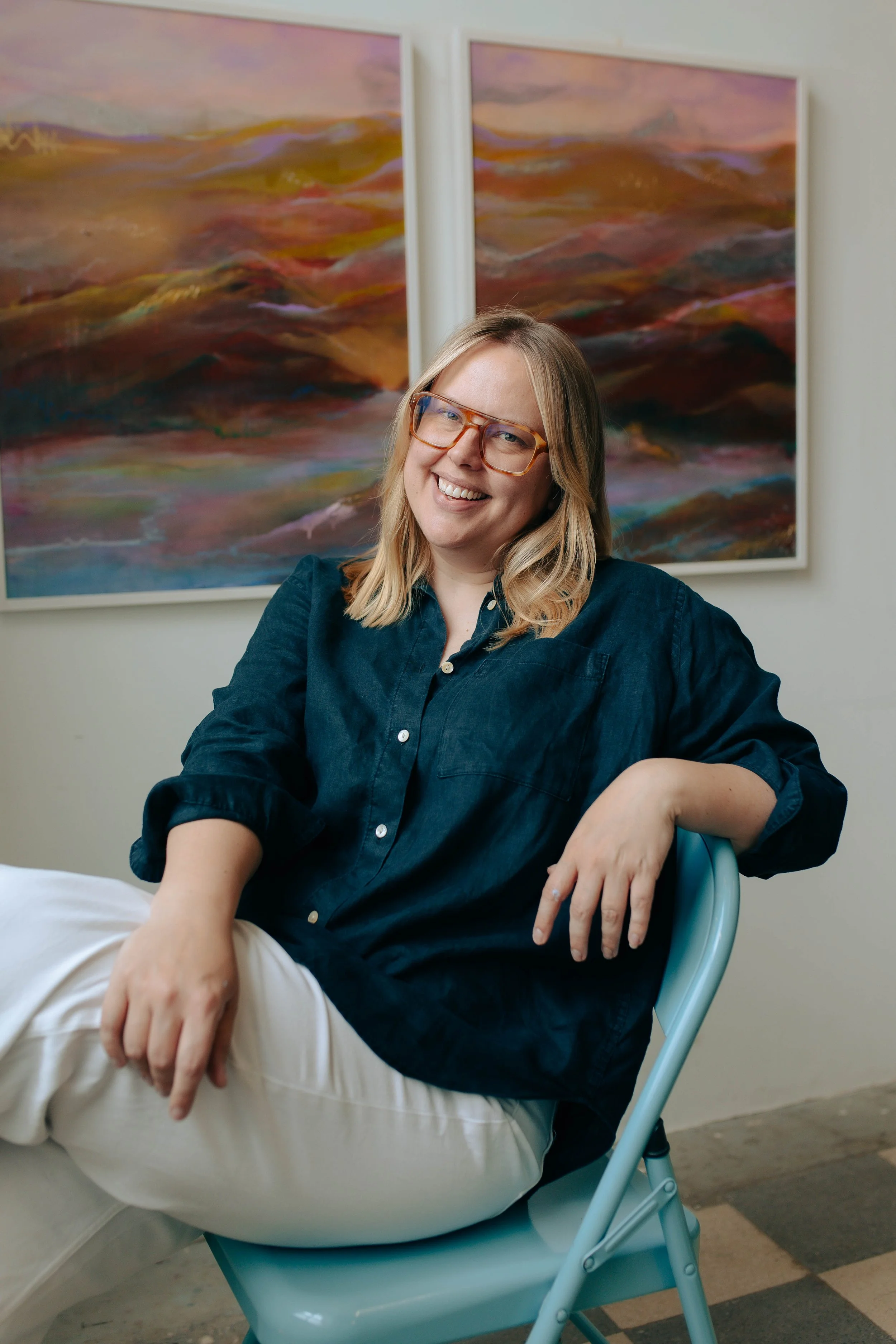 A white woman with short blonde hair, glasses, and a navy blue shirt sitting in a light blue chair in front of abstract landscape paintings, smiling at the camera.