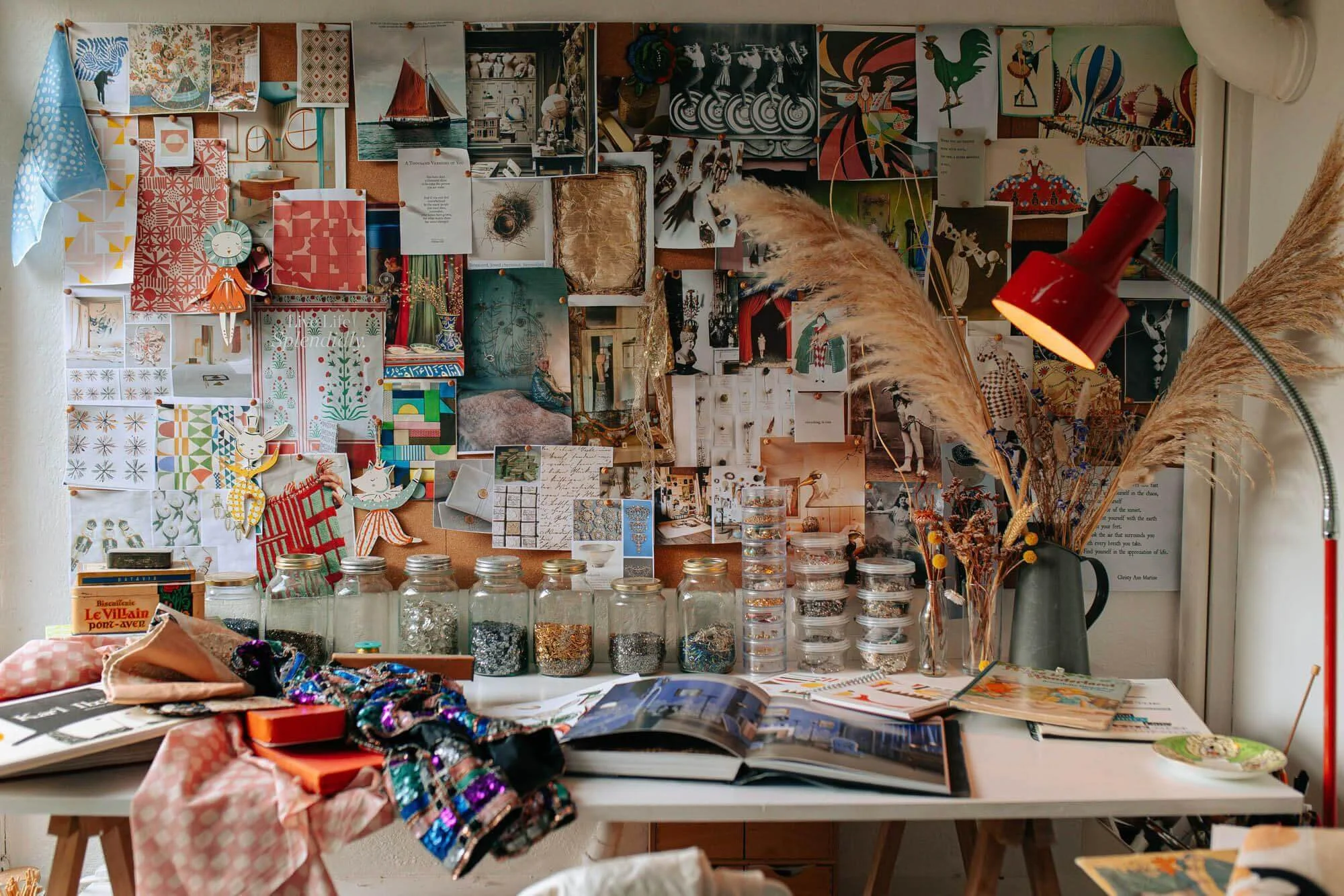 A creative workspace with a bulletin board filled with photos, artwork, and notes, a table with open books, jars of beads, fabric, and crafting supplies, and a vase with dried pampas grass and flowers.