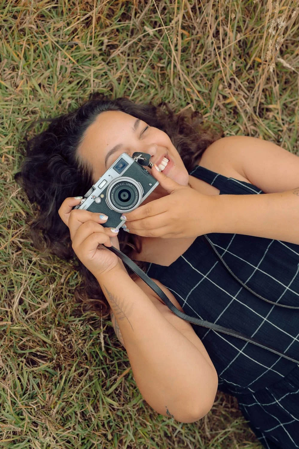A woman with curly dark hair lying on grass, smiling, holding a vintage camera to her face.