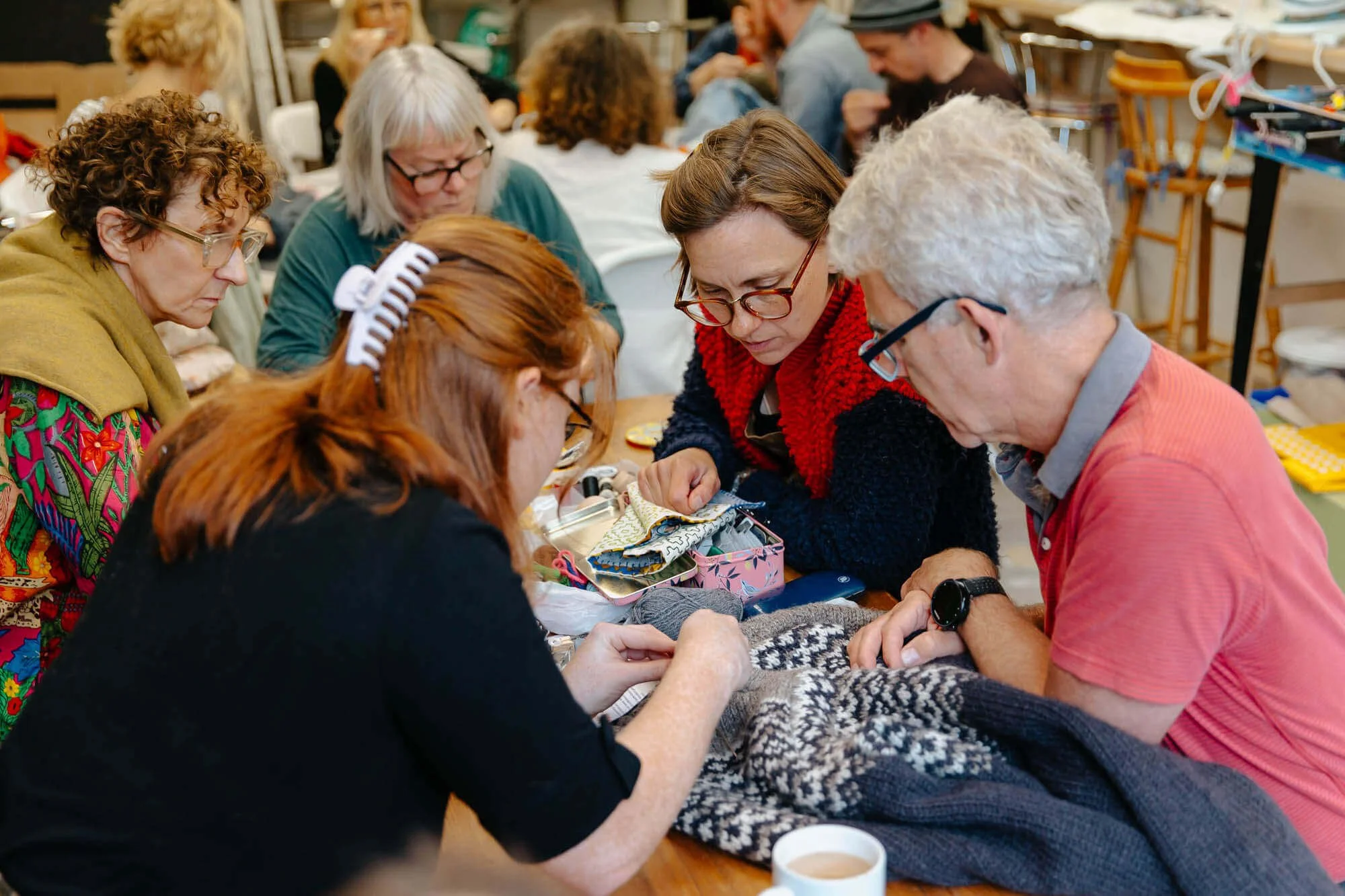 A group of people gathered around a table, working together on a knitting project in a craft or community center, by Celie Nigoumi, Brand and Events Photographer