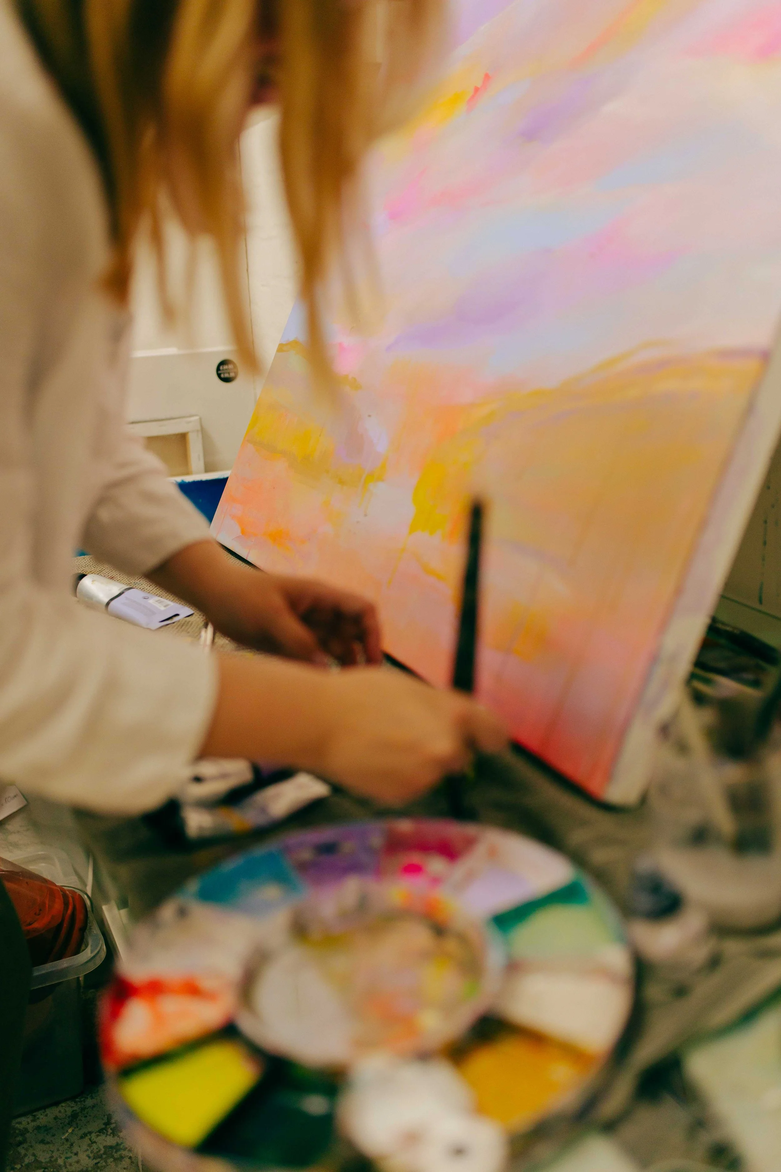 Close-up of an artist's workspace showing a partially visible watercolor painting with pink, purple, and yellow hues, surrounded by various art supplies, including paints and brushes.
