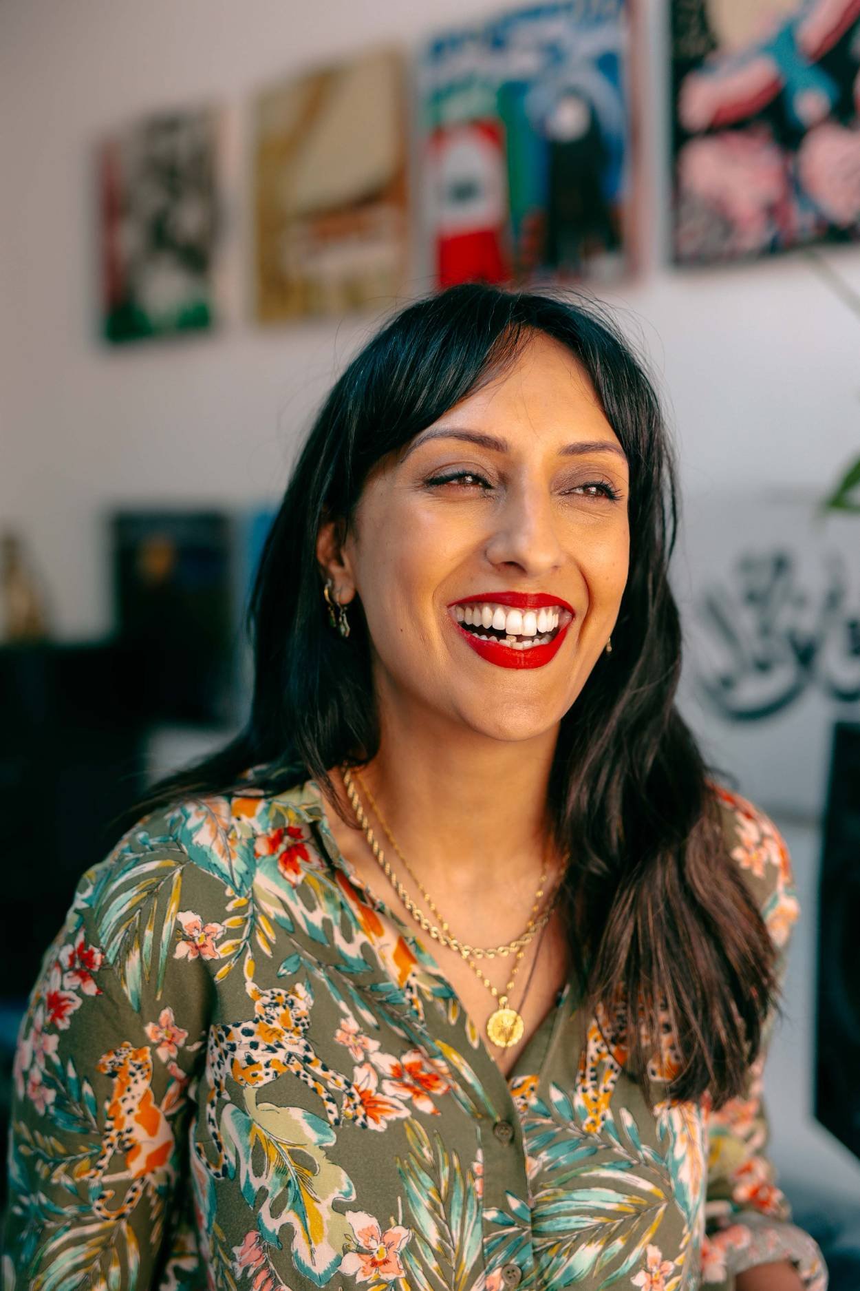 A woman with dark hair, wearing red lipstick, gold jewelry, and a floral shirt, smiling in an indoor setting with artwork in the background.