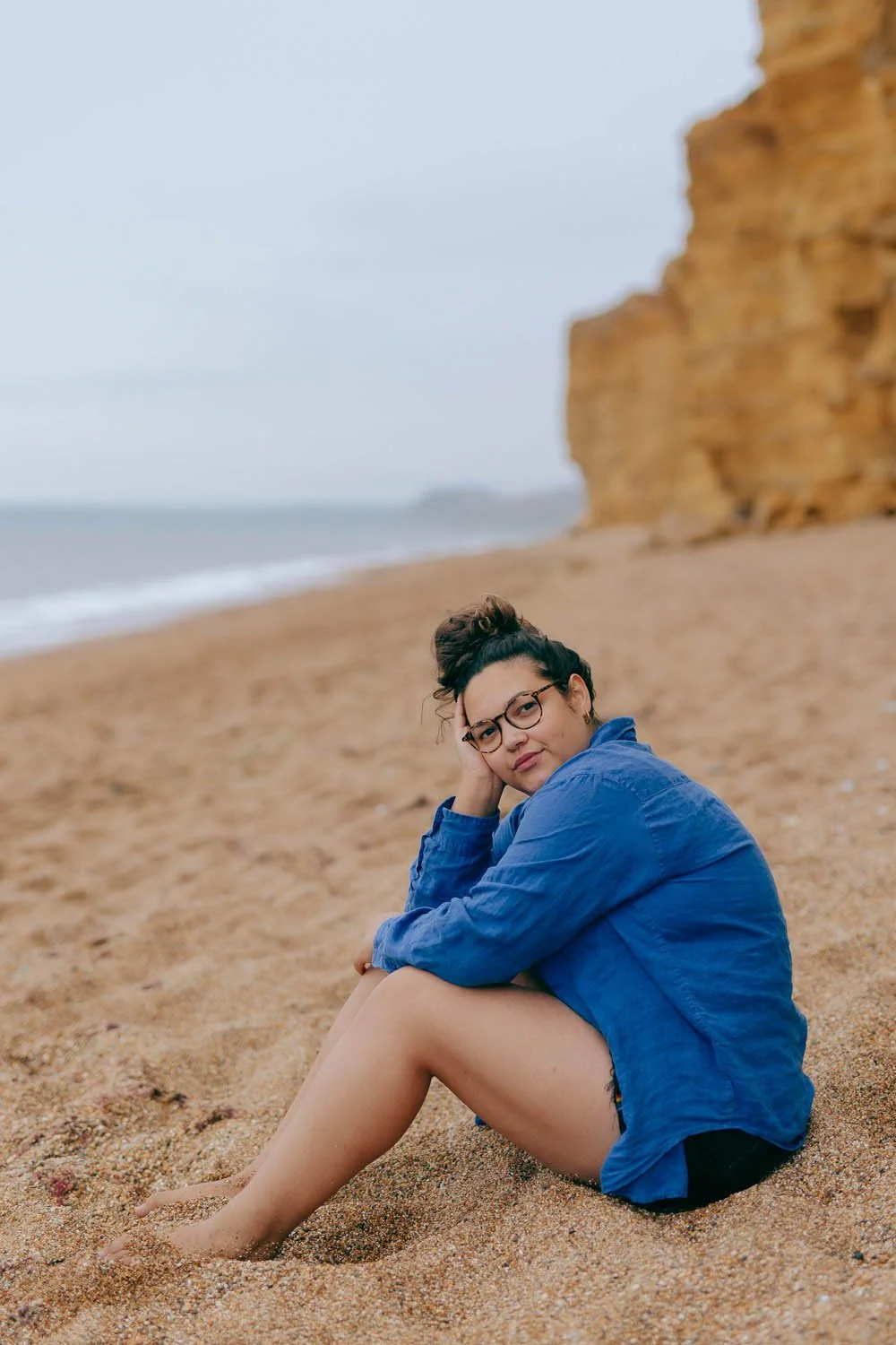 Woman with curly hair and glasses, in a blue shirt, sitting on a beach