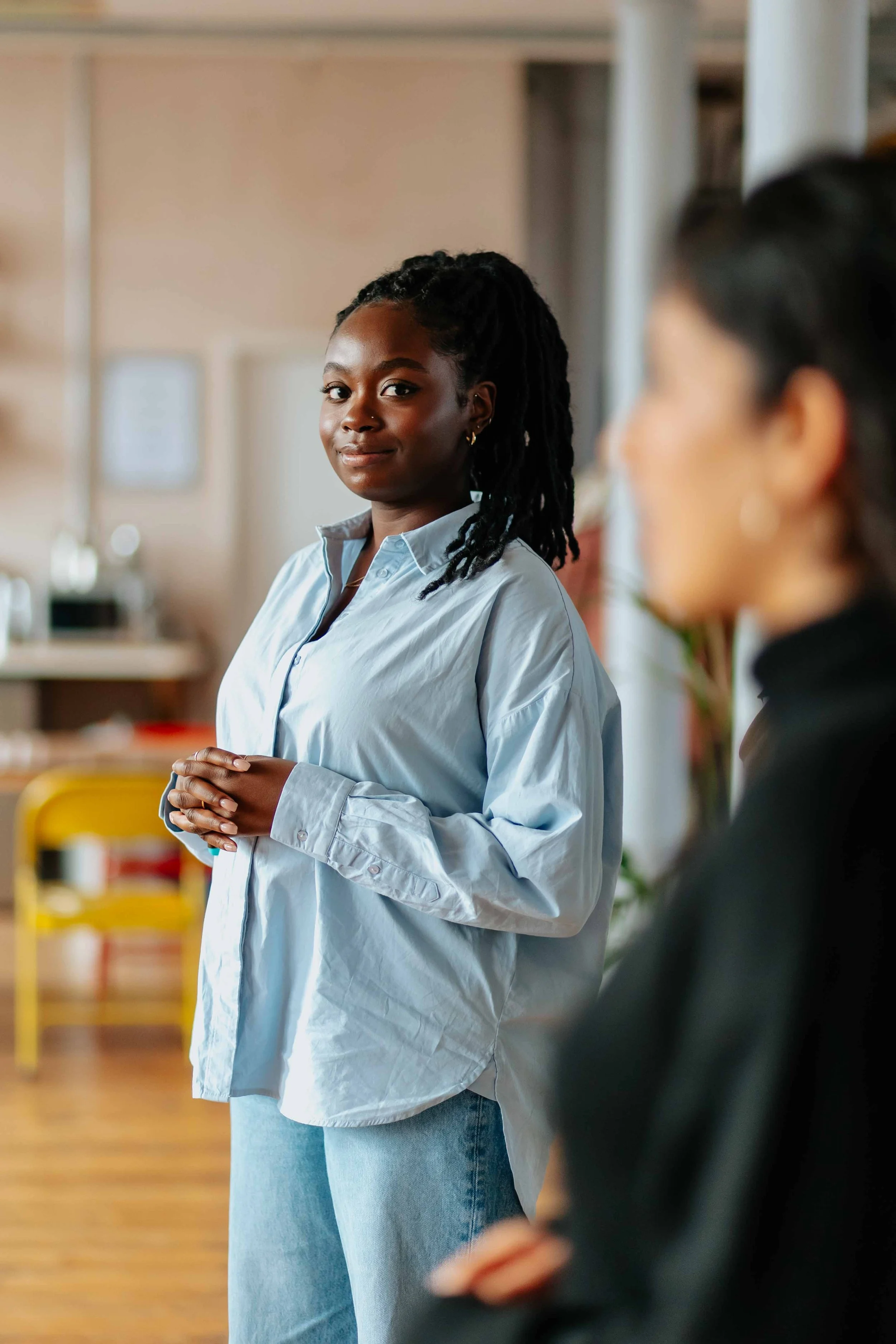 A young Black woman with dreadlocks wears a light blue shirt and jeans, standing with her hands clasped, smiling confidently indoors with blurred background.