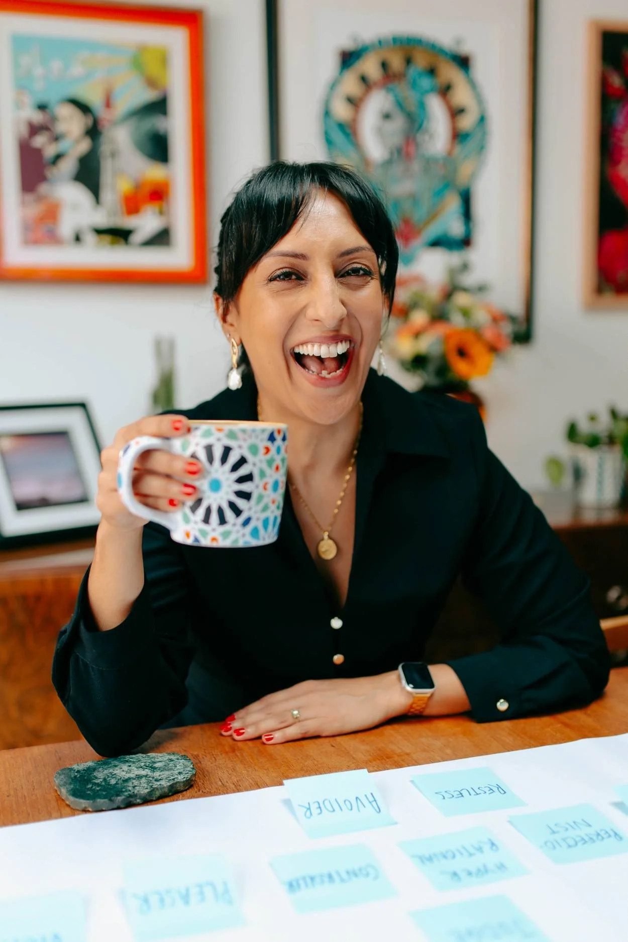A woman smiling and holding a colorful mug, sitting at a table with notes and a rock on it in an art-filled room.