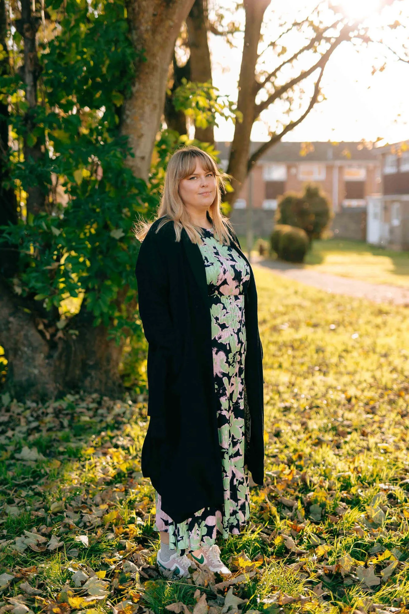 A woman standing outdoors in a park-like setting with trees and grass, wearing a patterned dress, black coat, and sneakers during sunset.