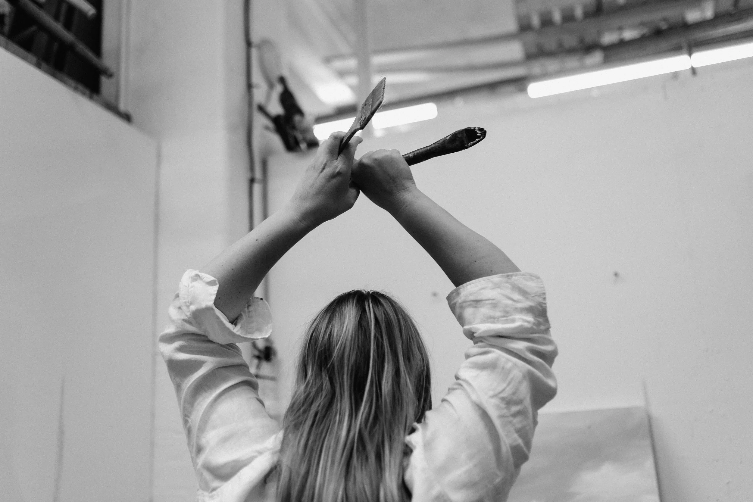 A black and white photograph of a person with long hair wearing a white shirt is holding a paintbrush and a palette, raised above their head, in a room with fluorescent lights.