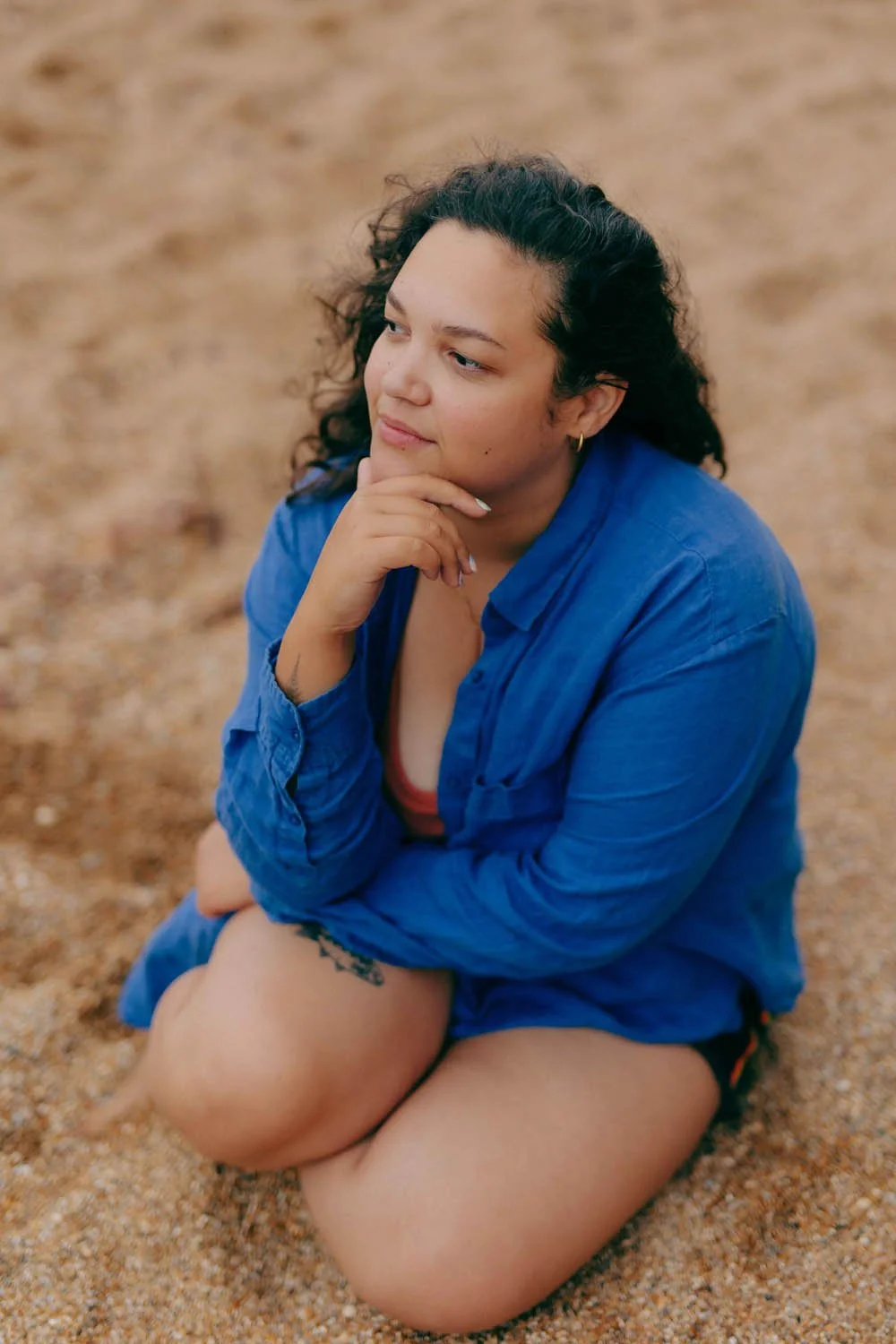 Woman with curly hair, in a blue shirt, sitting on a beach resting her chin on her hand