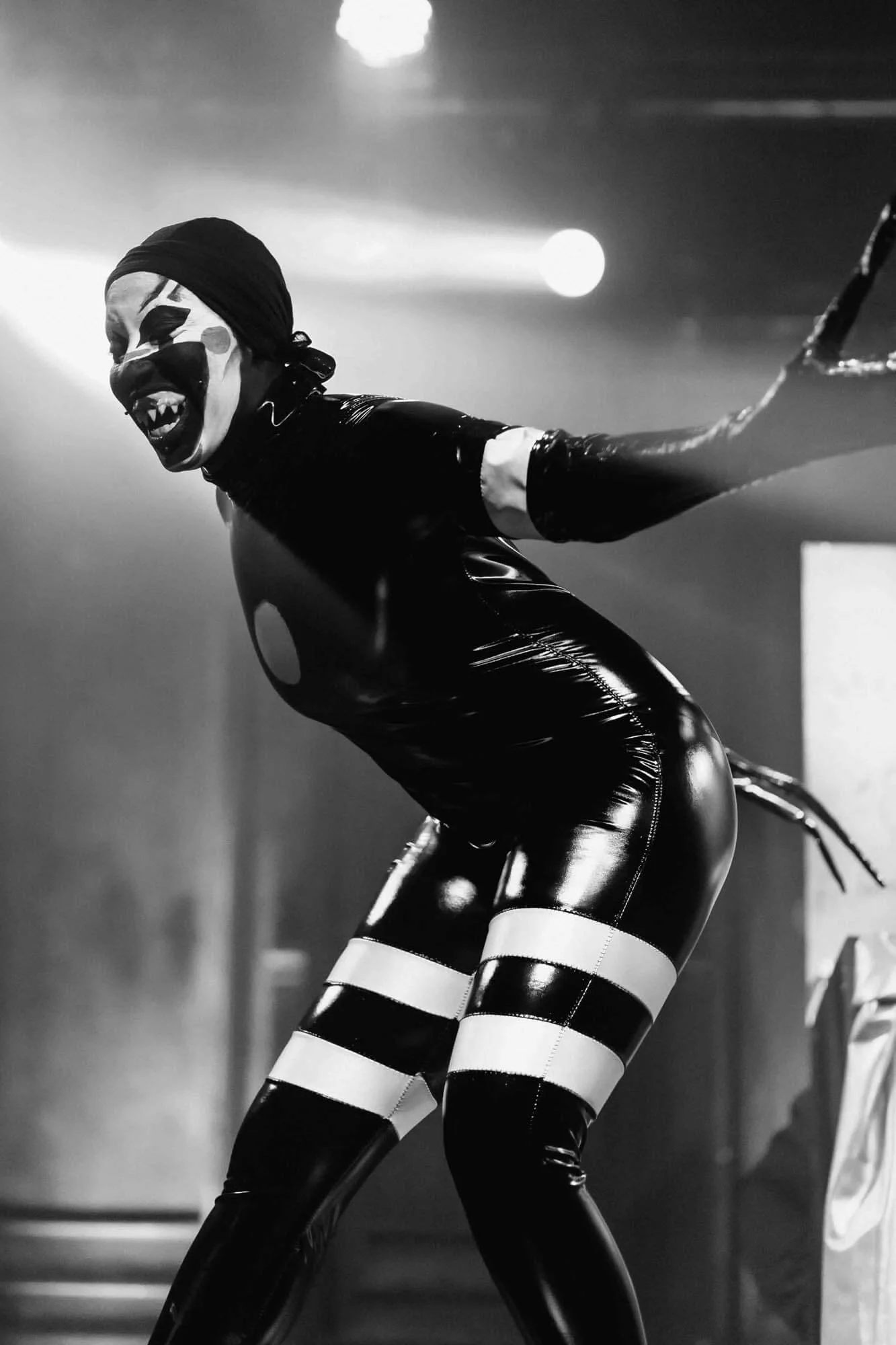 A person dressed in a shiny black latex bodysuit and striped thigh-high stockings, wearing clown makeup and a headscarf, performing on stage with an energetic and mischievous expression.