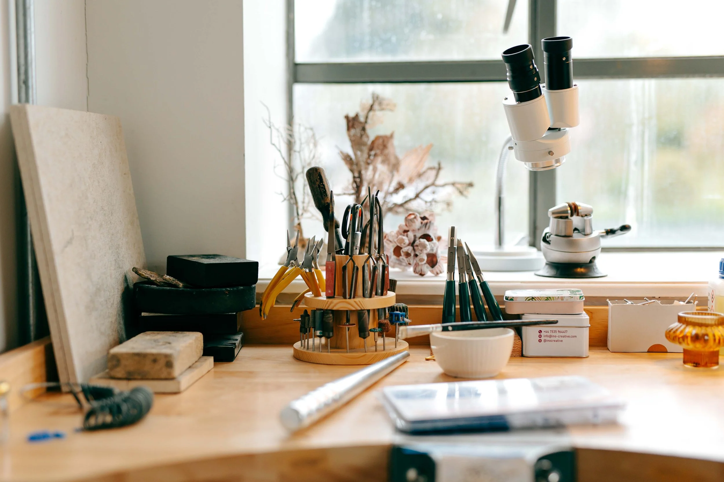 A workbench in a workshop with various tools including screwdrivers and pliers, a microscope, and art supplies, with a window in the background.