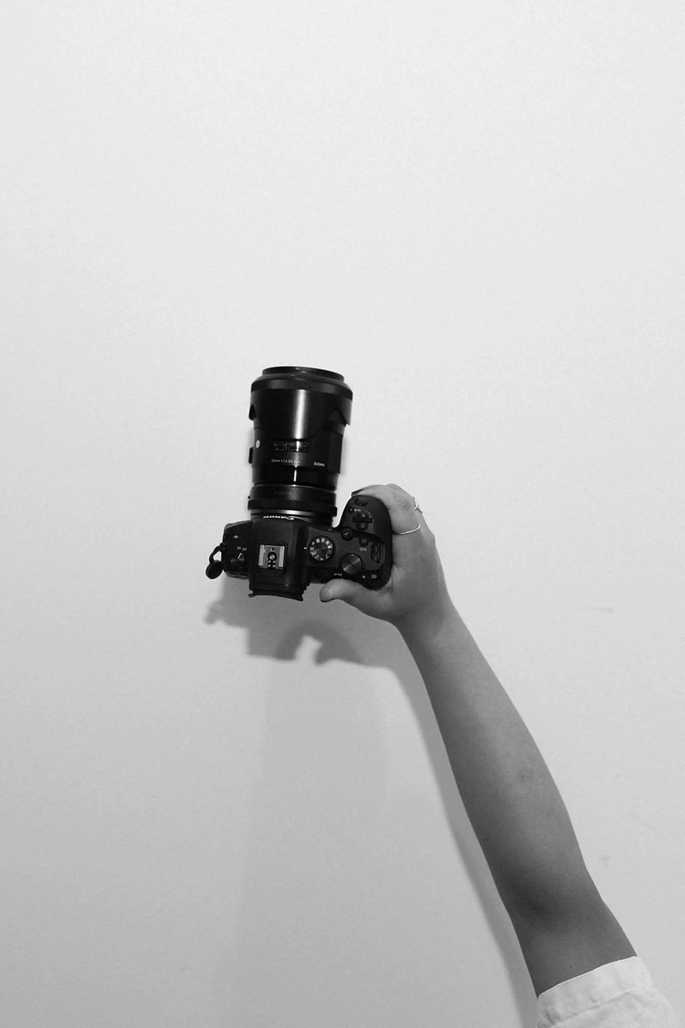 A hand holding a DSLR camera with a long zoom lens against a plain white wall.