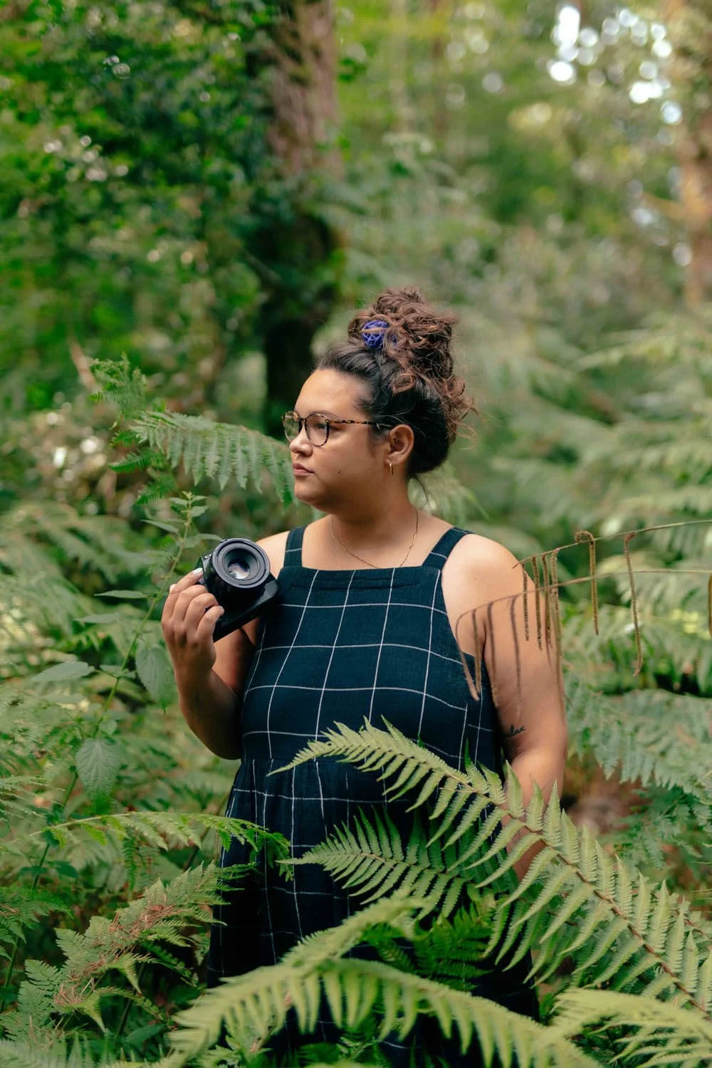 A woman with glasses and curly hair tied up, wearing a black dress with white grid pattern, holding a camera in a lush green forest. Celie Nigoumi Lifestyle, Editorial, Events Brand and Portraits Photographer in Frome, Somerset