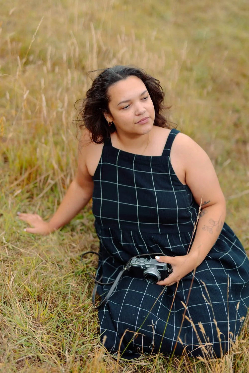 Young woman with curly hair sat in a meadow, in a checkered dress.
