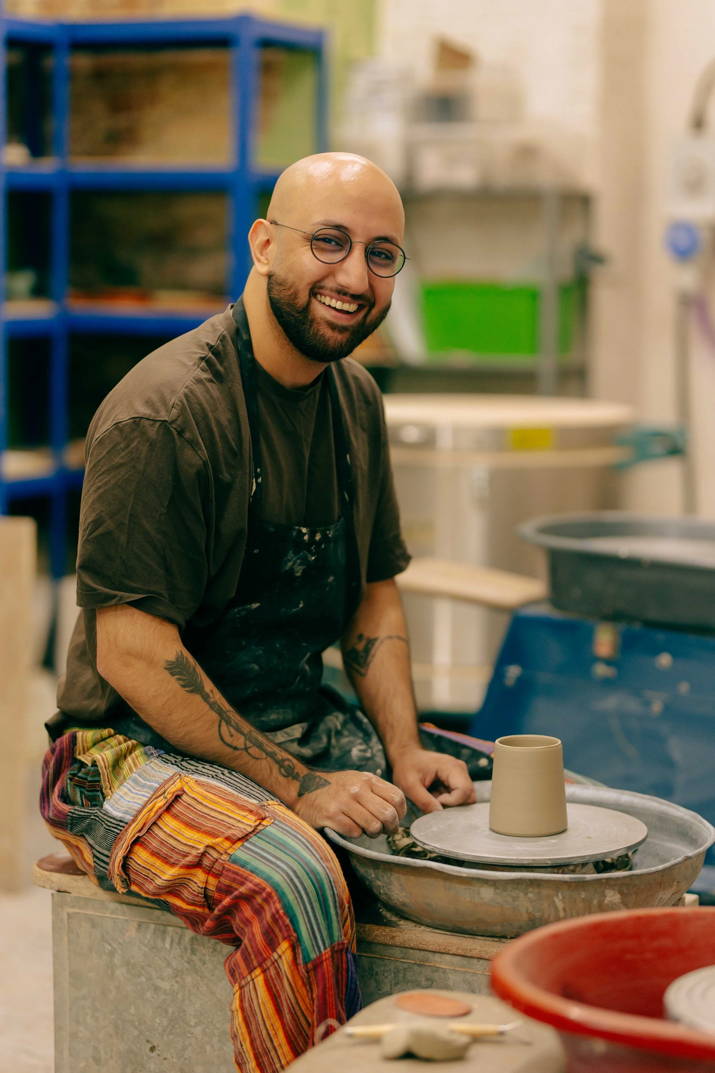 A smiling bald man with glasses and a beard, wearing a dark shirt and colorful striped pants, sitting at a pottery wheel with a clay cup in progress, in a ceramics studio filled with shelves holding various pottery supplies.