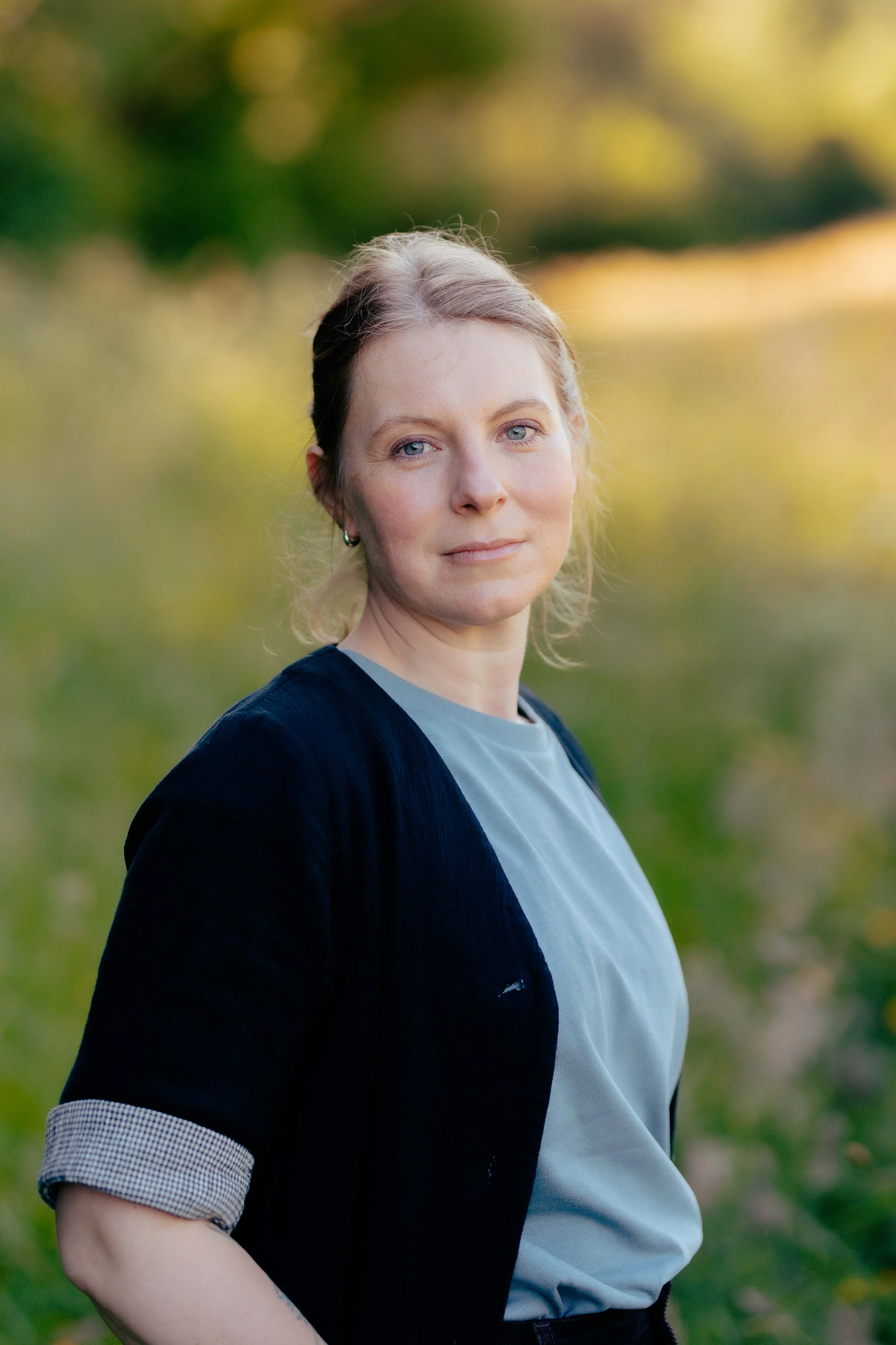 A white woman with blue eyes and light brown hair pulled back, wearing a light blue shirt and a dark jacket with rolled sleeves, standing outdoors against a blurred background of trees and greenery.