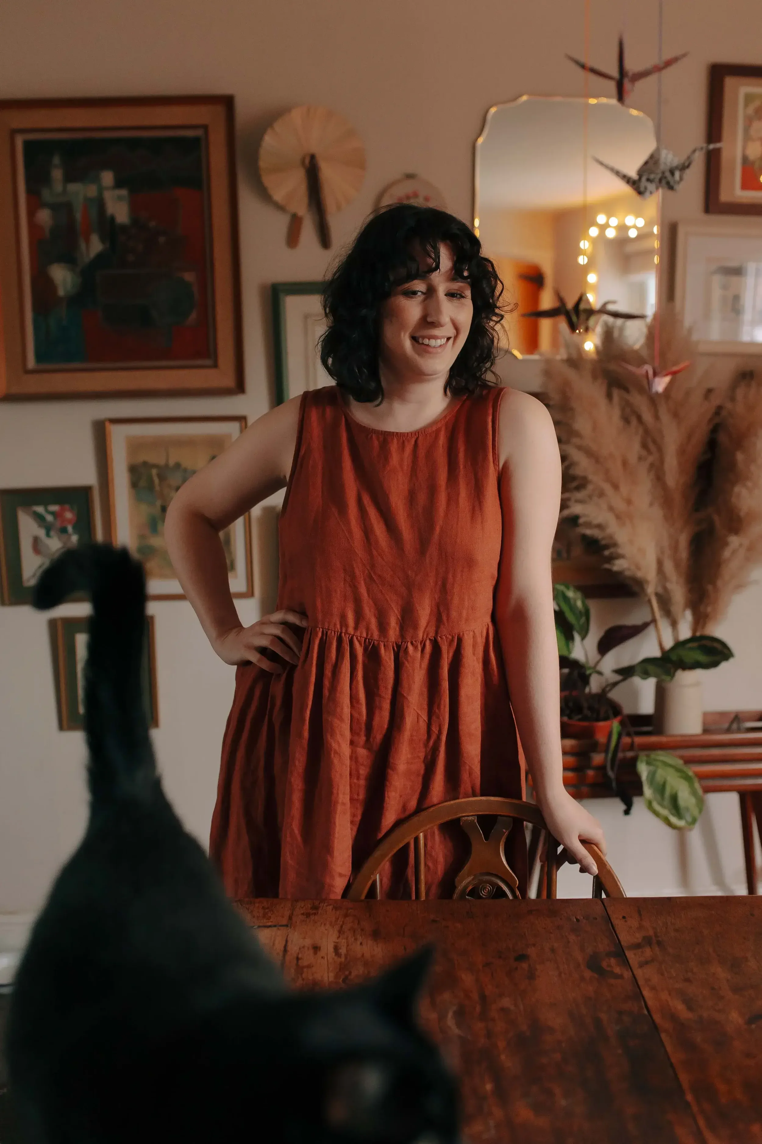 A woman in an orange sleeveless dress standing behind a wooden table smiling at a black cat in the foreground, with a decorated wall and hanging plants in the background.