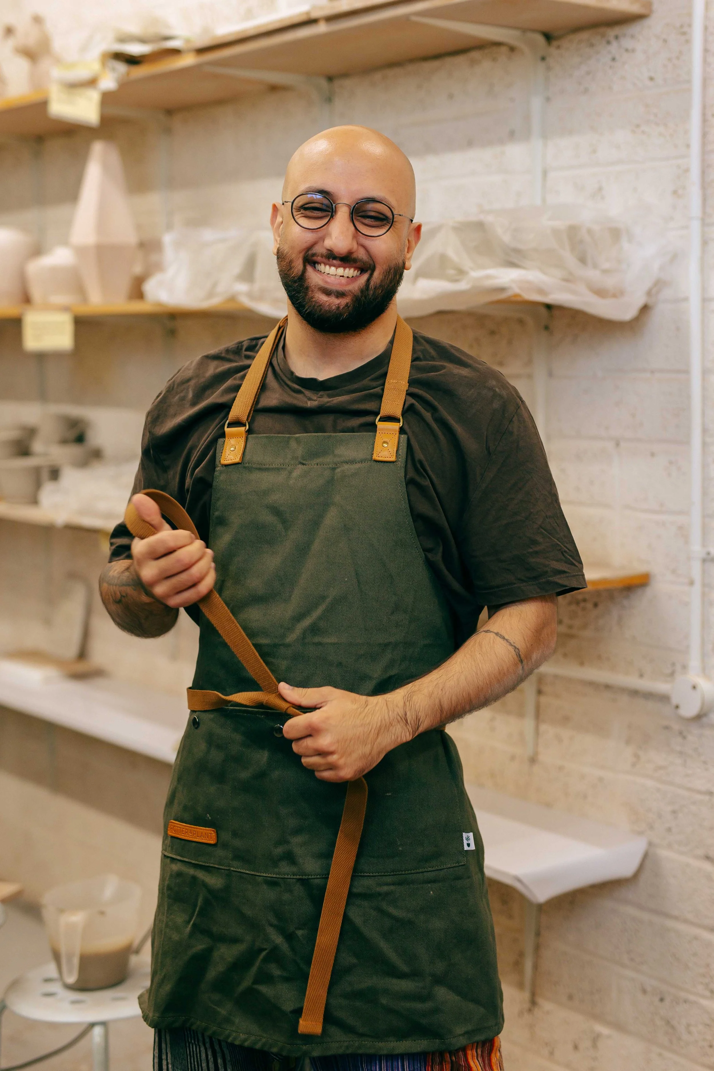 A smiling man with glasses, a beard, and a shaved head wearing a dark shirt and an apron, standing in a brick-walled room with shelves and packaging materials in the background.