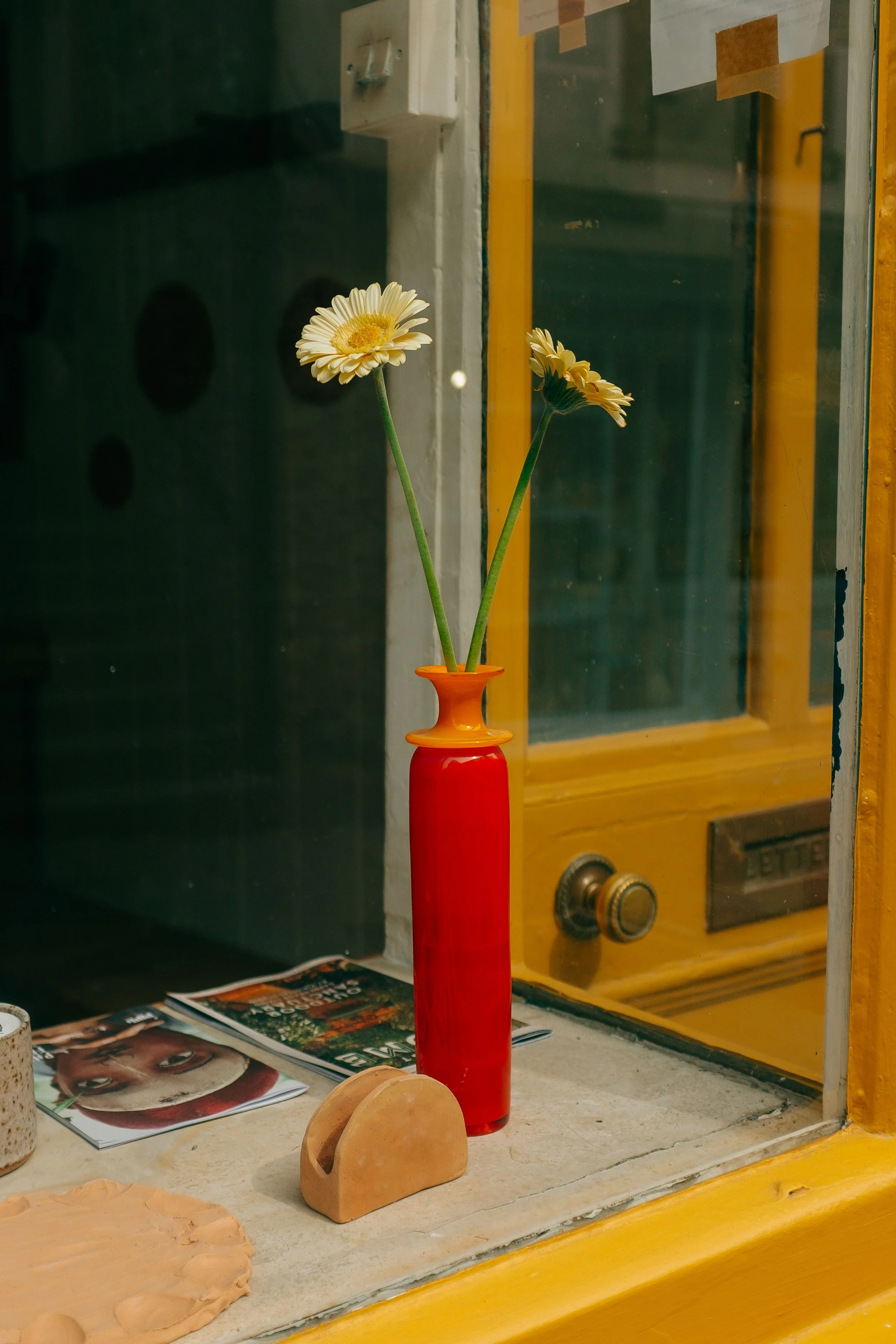 A tall red vase with two yellow daisies on a picture window sill, with magazines and a small ceramic object nearby. By Celie Nigoumi, Lifestyle Photographer
