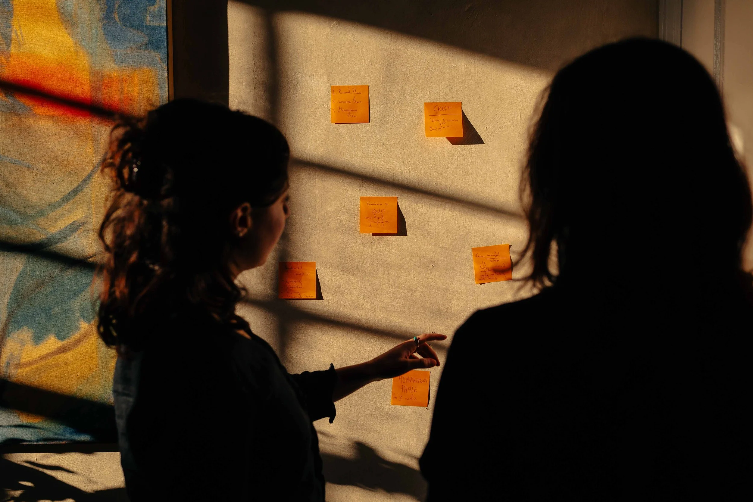 Two women standing in front of a wall with orange sticky notes, with shadows cast across the scene.