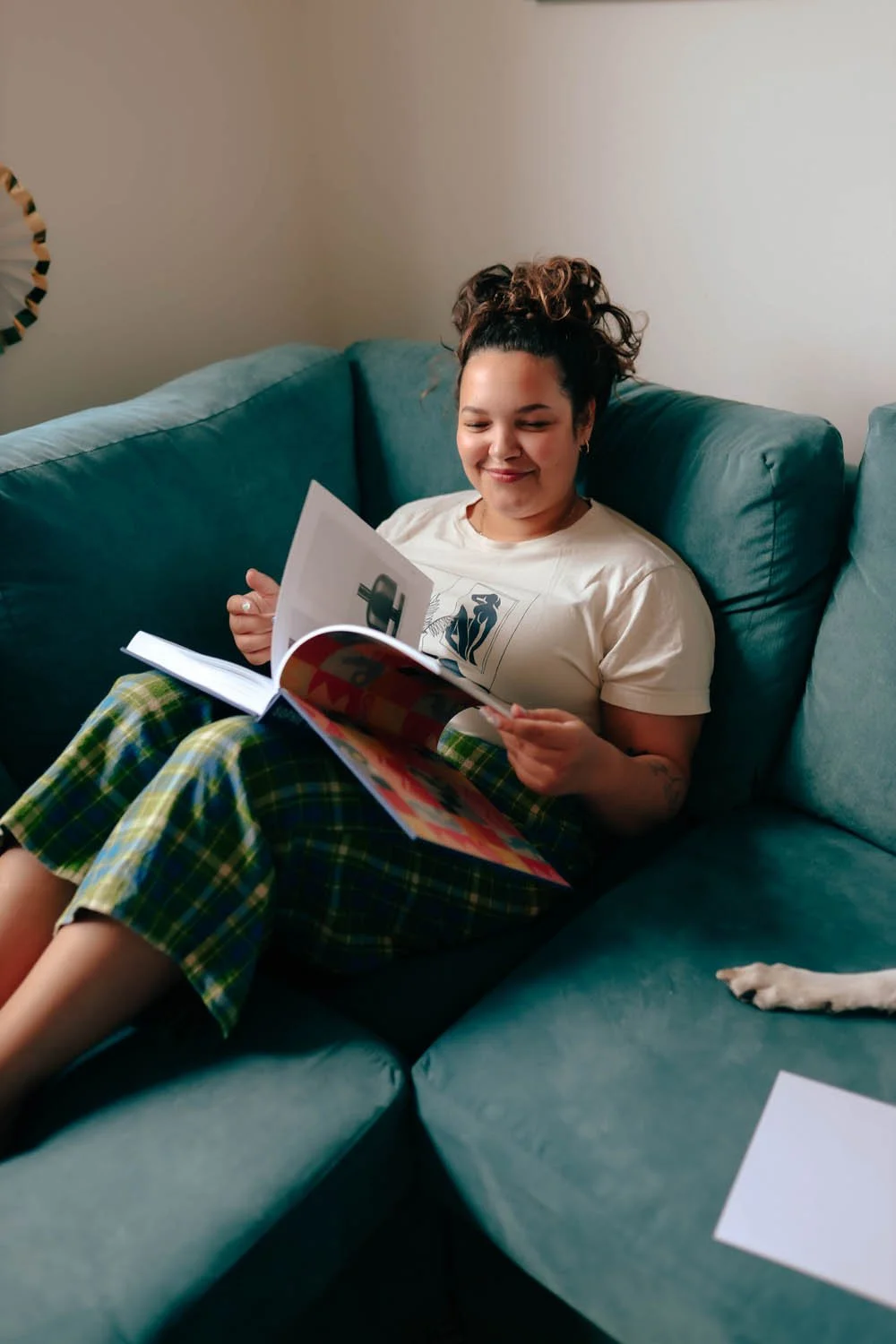 Woman with curly hair smiling, sitting on a teal sofa, reading a magazine, wearing a cream T-shirt and green plaid pants.
