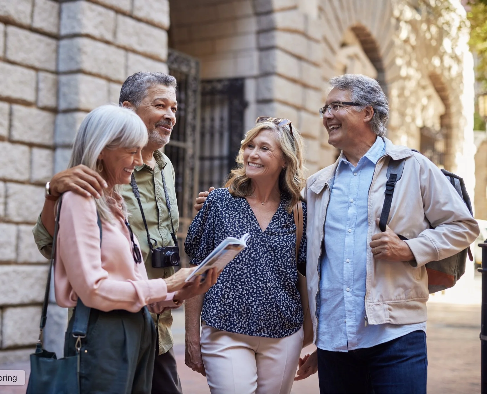 Four older adults smiling and chatting on a city street, one woman reading a guidebook, in casual outdoor clothing.
