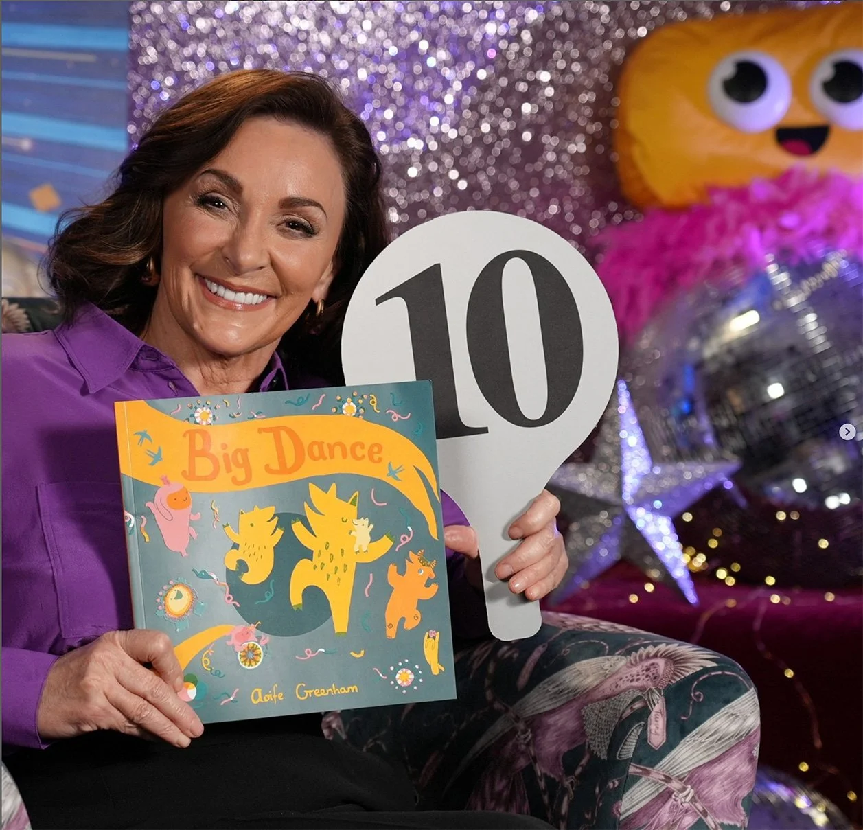 A woman, Shirley Ballas, sits on a couch, smiling and holding the picturebook Big Dance in one hand, and a judges scorecard 10 in the other. She is on the colourful and sparkly set of the ceebeebies bedtime story show. 