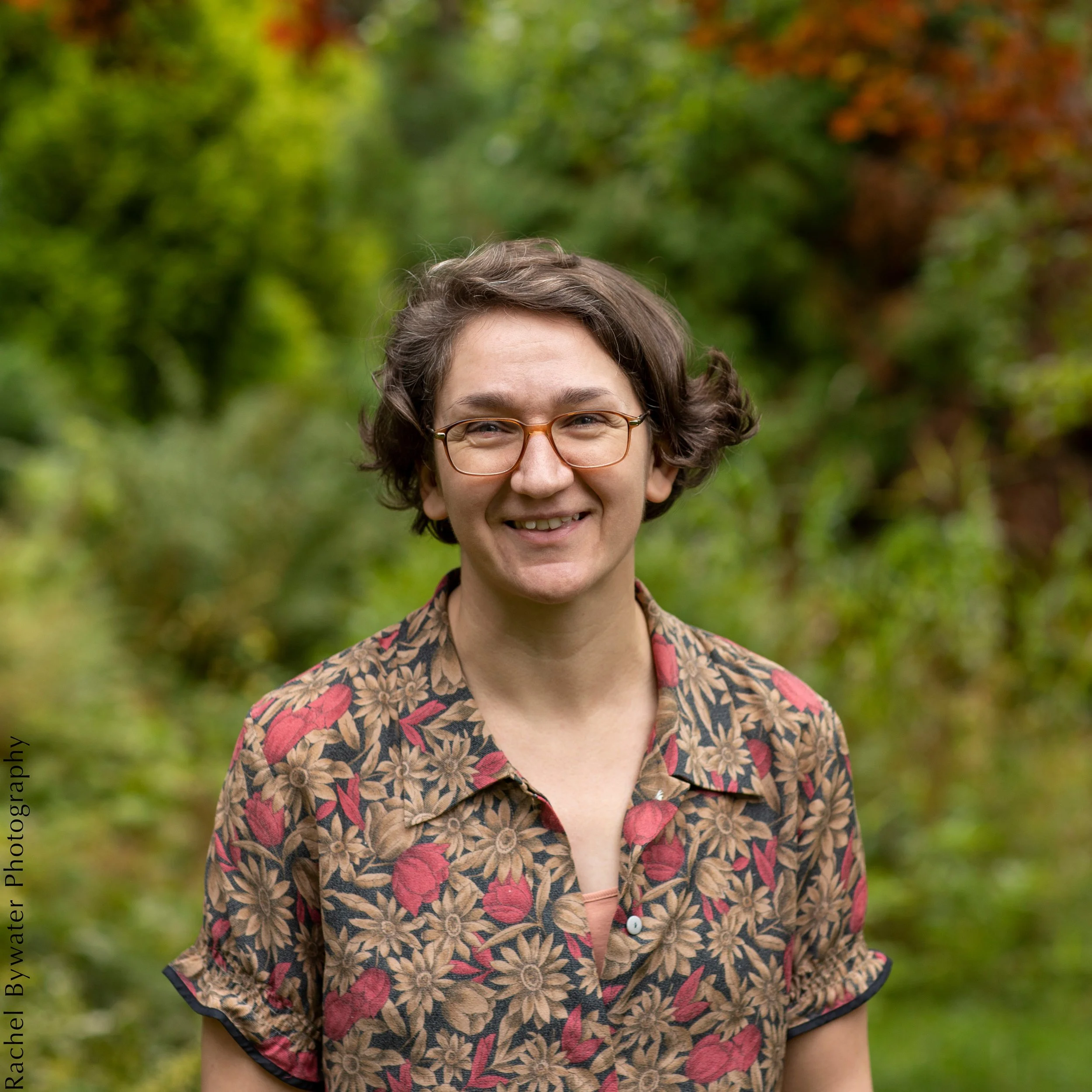 The illustrator Aoife Greenham, a woman with short brown hair and glasses, uncomfortable about having her photo taken, is posing in front of a lush green leafy background.