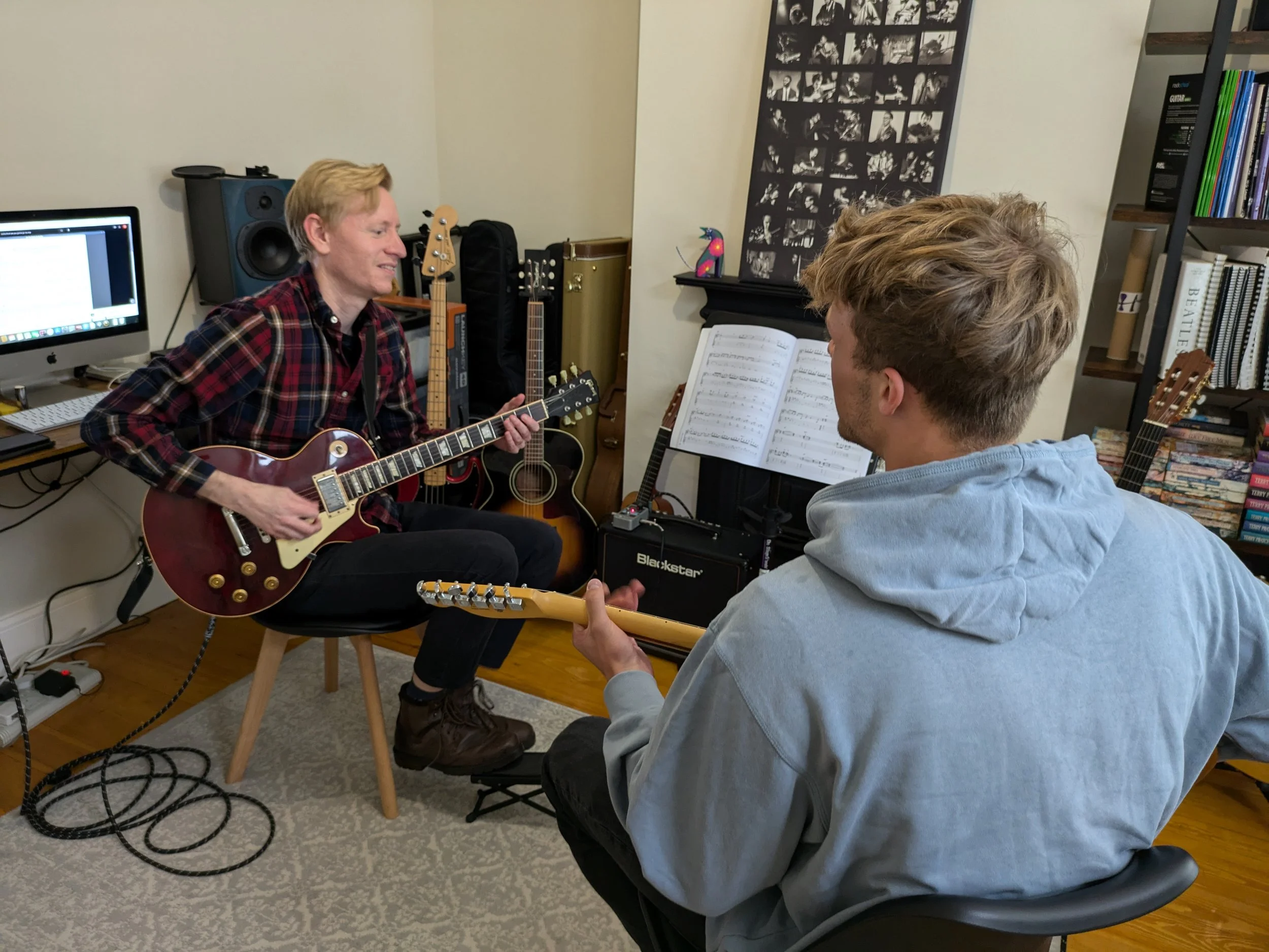 Two young men playing guitars in a music studio, with one sitting on a stool and the other in a chair, surrounded by musical equipment and books.