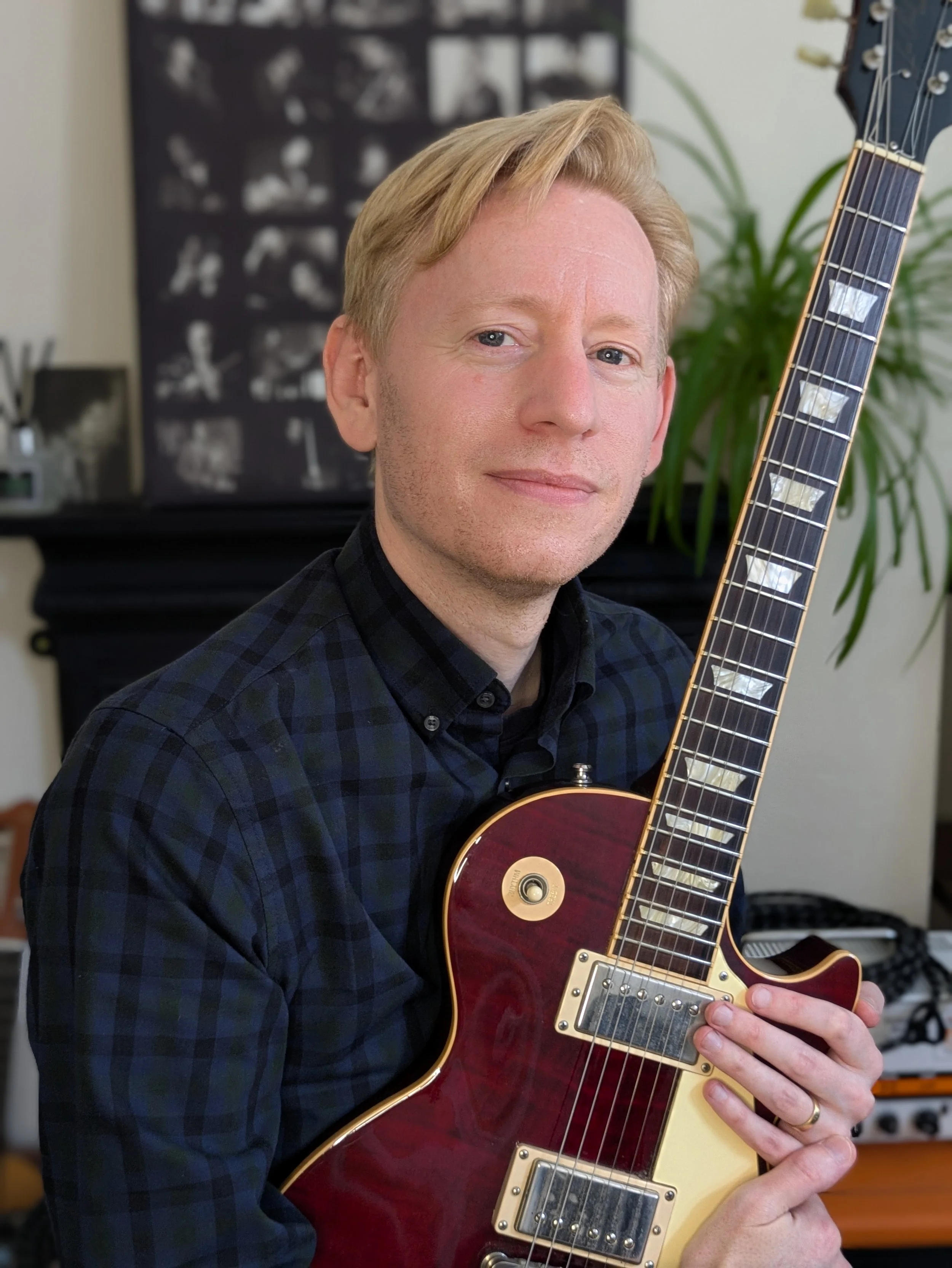 A man with blonde hair and a plaid shirt holding a red electric guitar with a black background and a plant in the background.