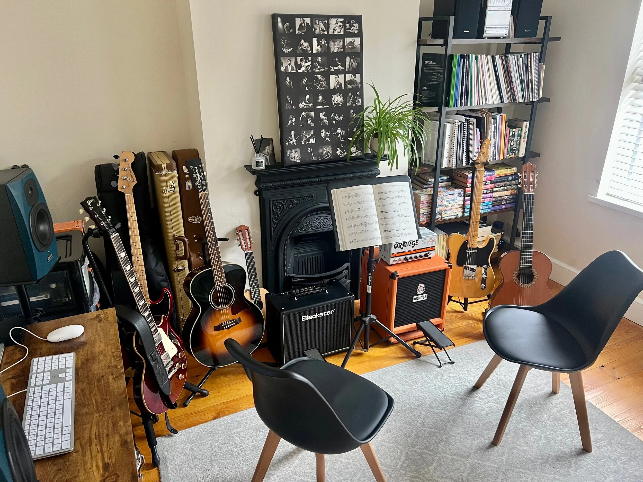Room with musical instruments including multiple guitars on stands, amplifiers, a music stand with sheet music, a black bookshelf filled with books and vinyl records, a computer monitor on a wooden desk, and a black chair in the foreground. There is a fireplace, a black framed photo collage, and a large window with blinds.