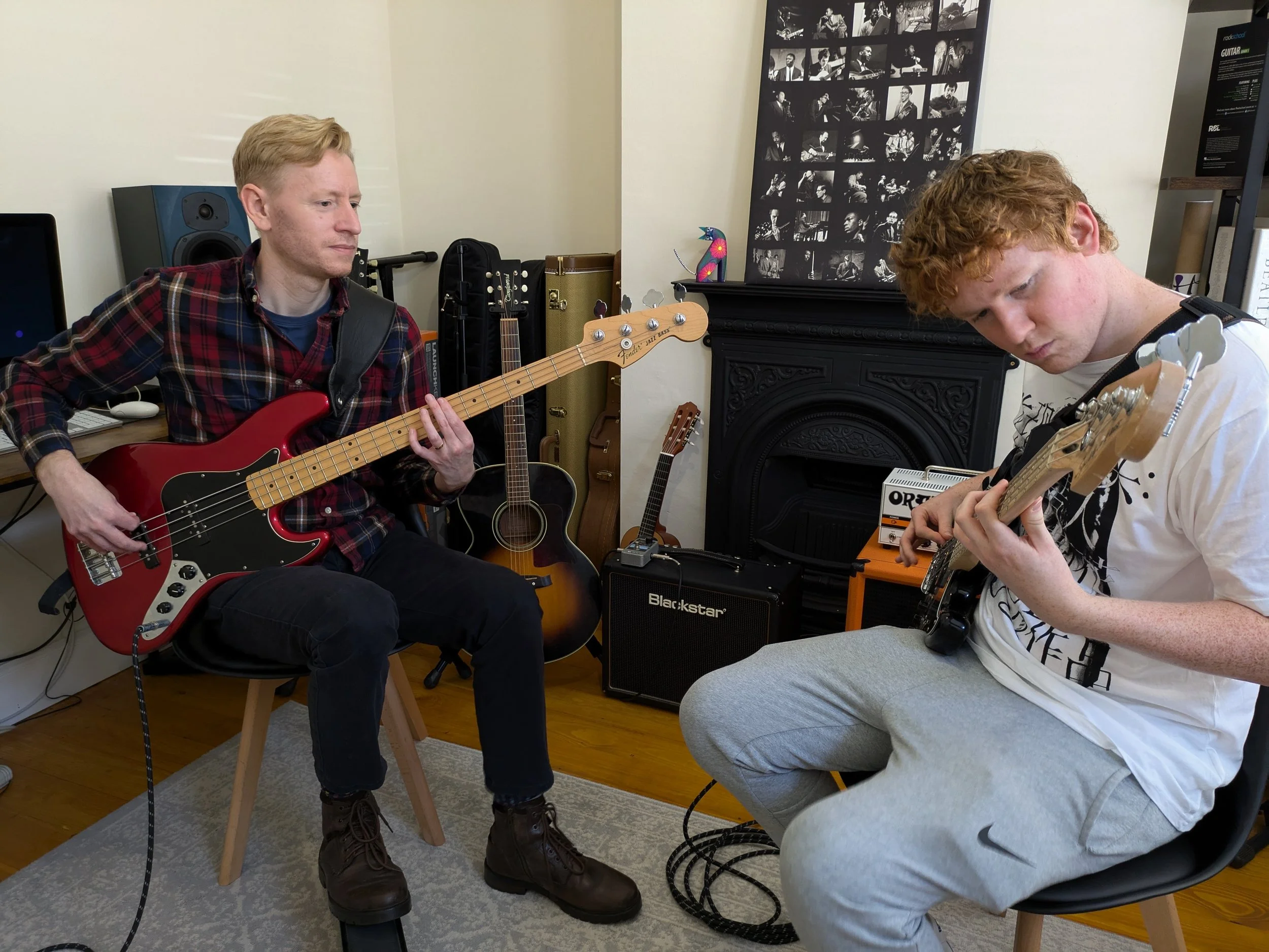 Two young men playing electric guitars in a room filled with musical equipment, including a bass guitar, acoustic guitar, small amplifier, and posters on the wall.