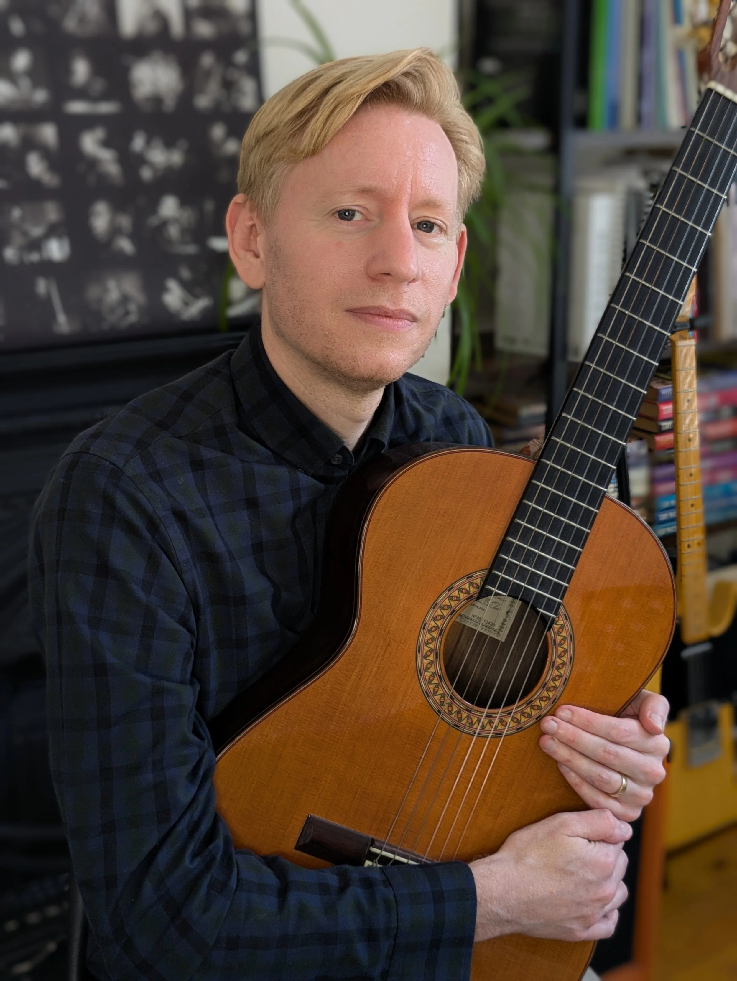 A man with blond hair wearing a dark plaid shirt, holding an acoustic guitar indoors with shelves of books.