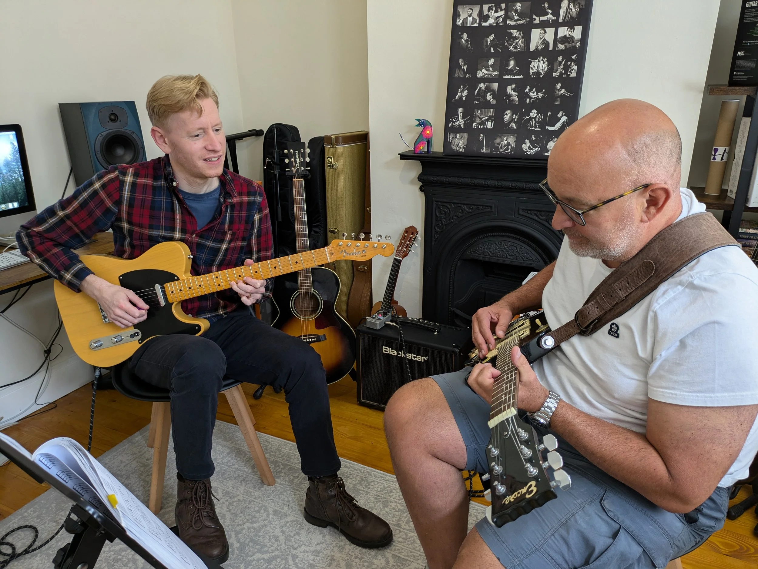 Two men playing acoustic guitars in a music room. One man is seated, smiling, with a yellow guitar. The other man is wearing glasses, a white t-shirt, and blue shorts, playing a black guitar. Several guitars and a music stand with sheet music are visible in the background.