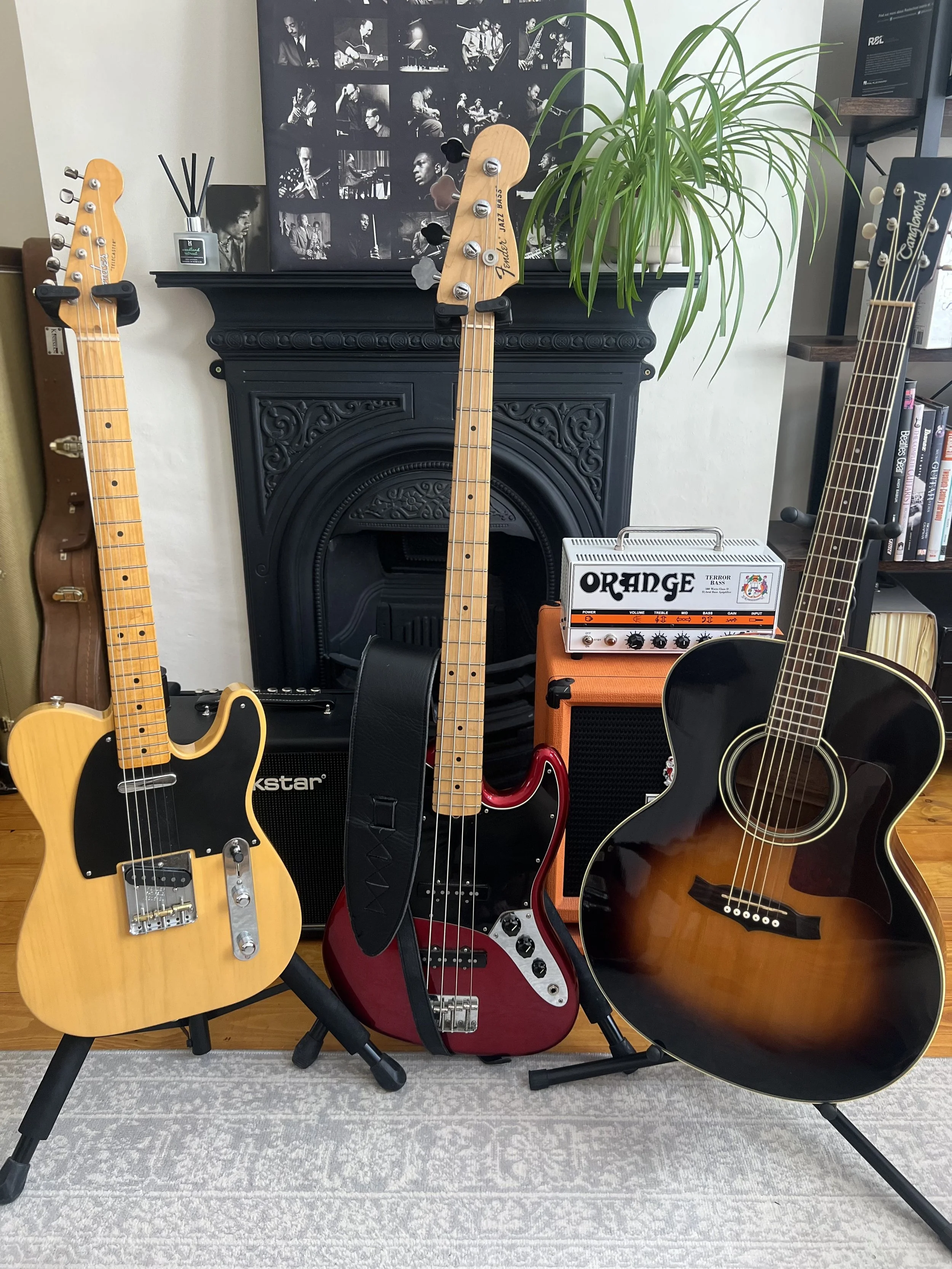 Collection of three electric and acoustic guitars on stands in front of a black ornate fireplace with a green plant on top. There is an orange amplifier and a small orange and white amplifier behind the guitars. A black and white photo collage hangs above the fireplace.