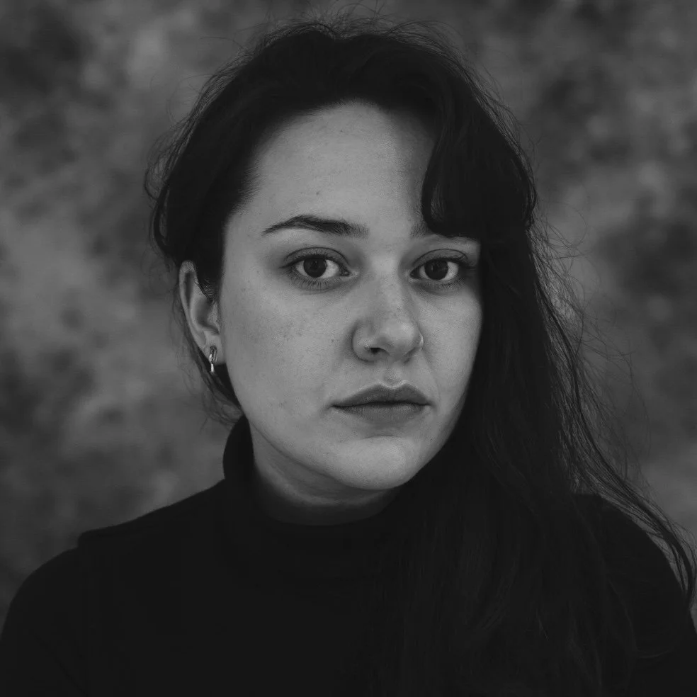 Black and white portrait of a young woman with long, dark hair, wearing a high-neck top, with neutral facial expression, and a blurred natural background.