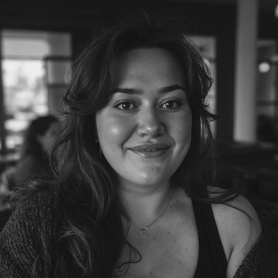 Black and white photo of a smiling woman with wavy hair, wearing a sleeveless top and a necklace, sitting indoors with a blurred background.