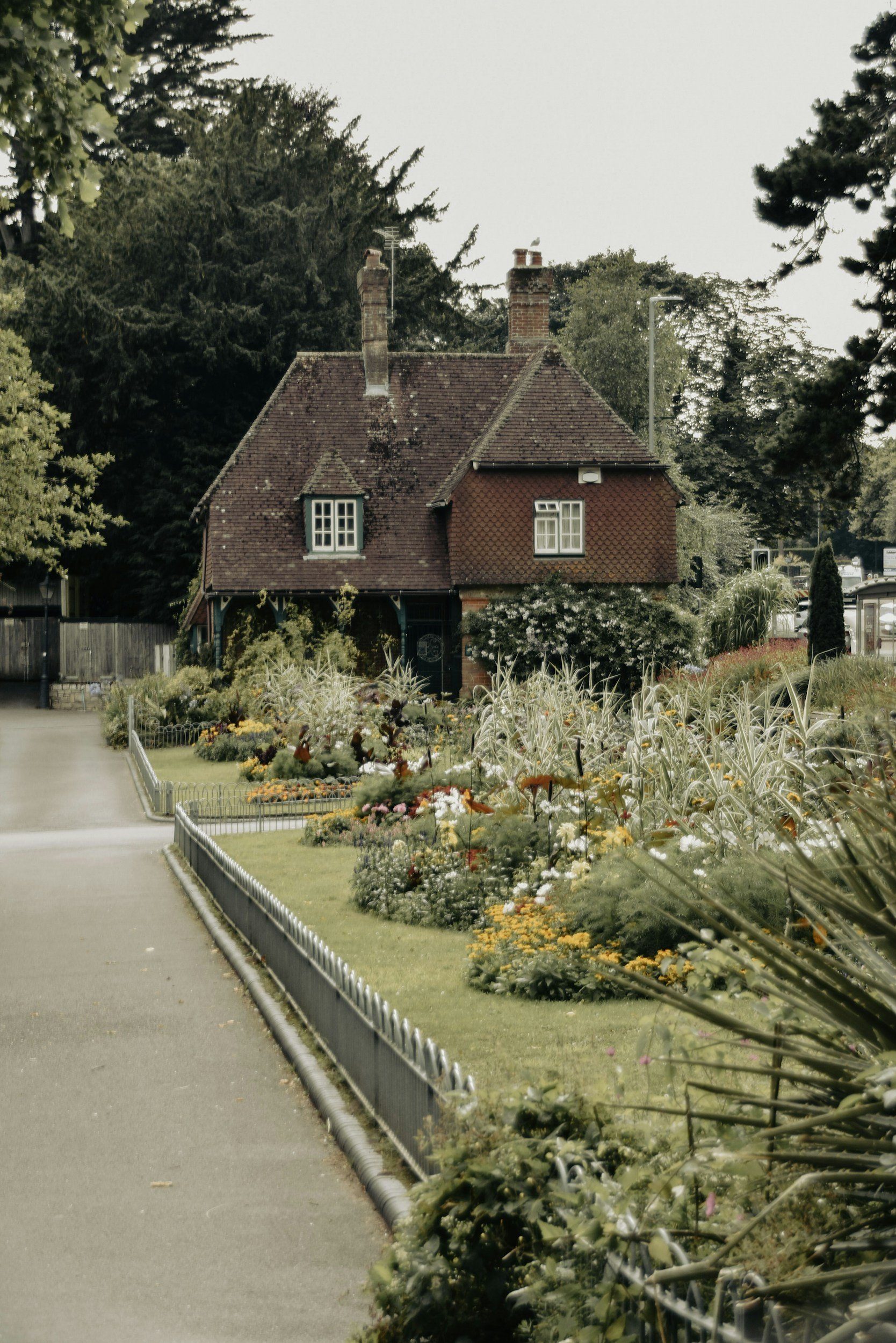 A house with a red tiled roof and chimney, surrounded by a garden with colorful flowers and greenery, along a pathway.