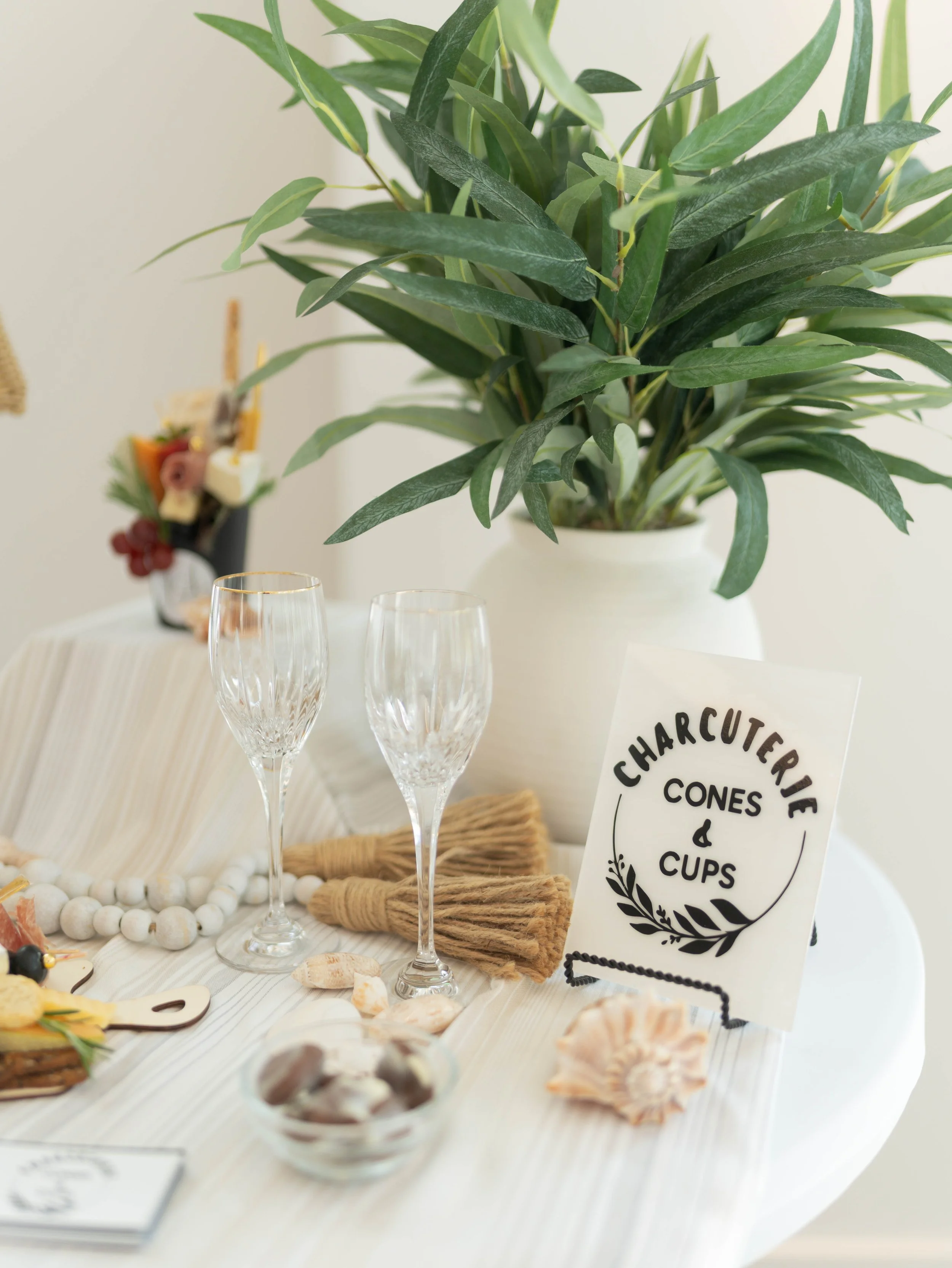A table decorated with seashells, glasses, and a sign reading "Charcuterie Cones & Cups." A large potted green plant is in the background.