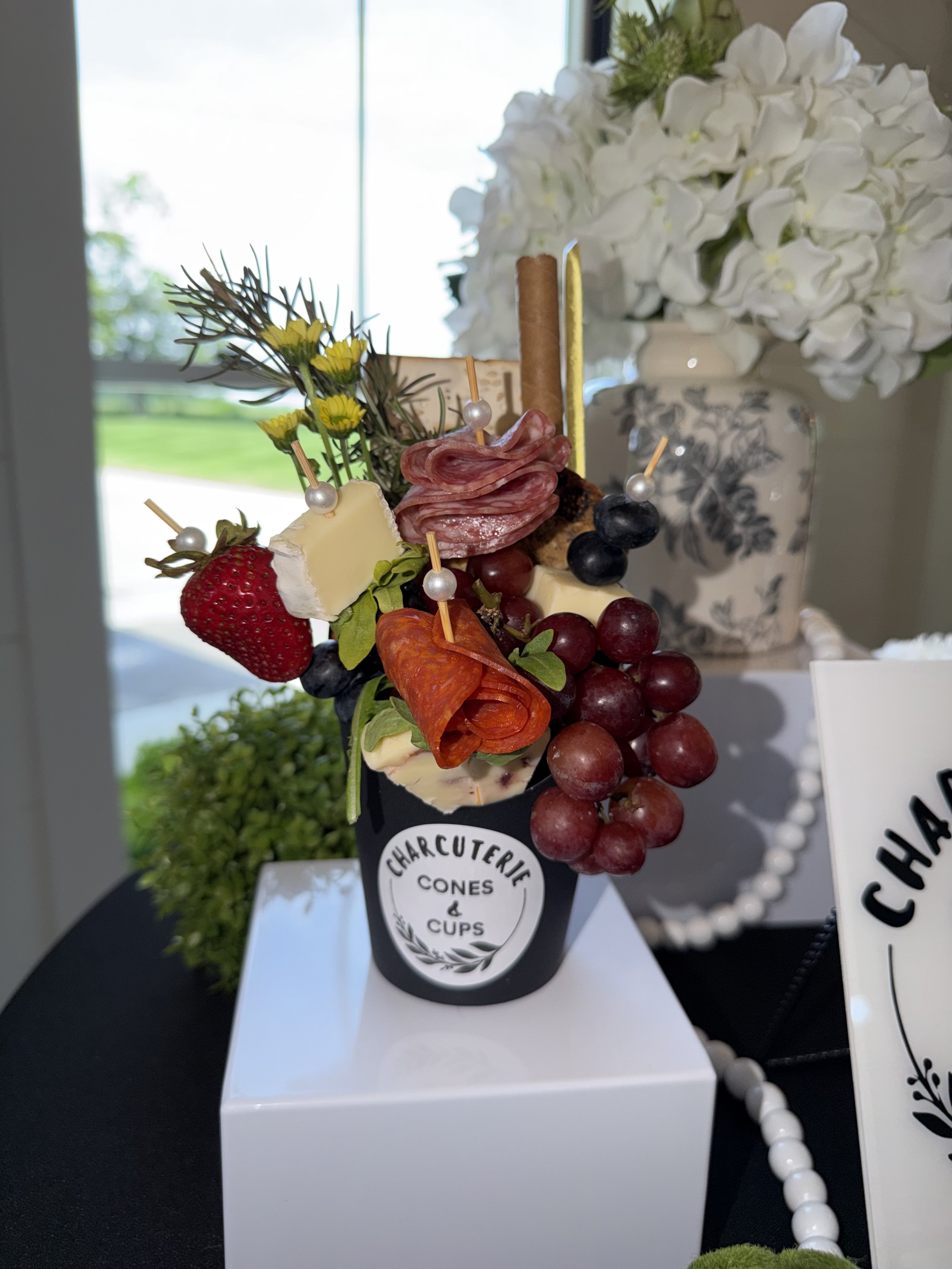 A bouquet with strawberries, cheese, grapes, salami, and decorative greenery on a black and white stand with a sign that reads "Charcuterie Cones & Cups"