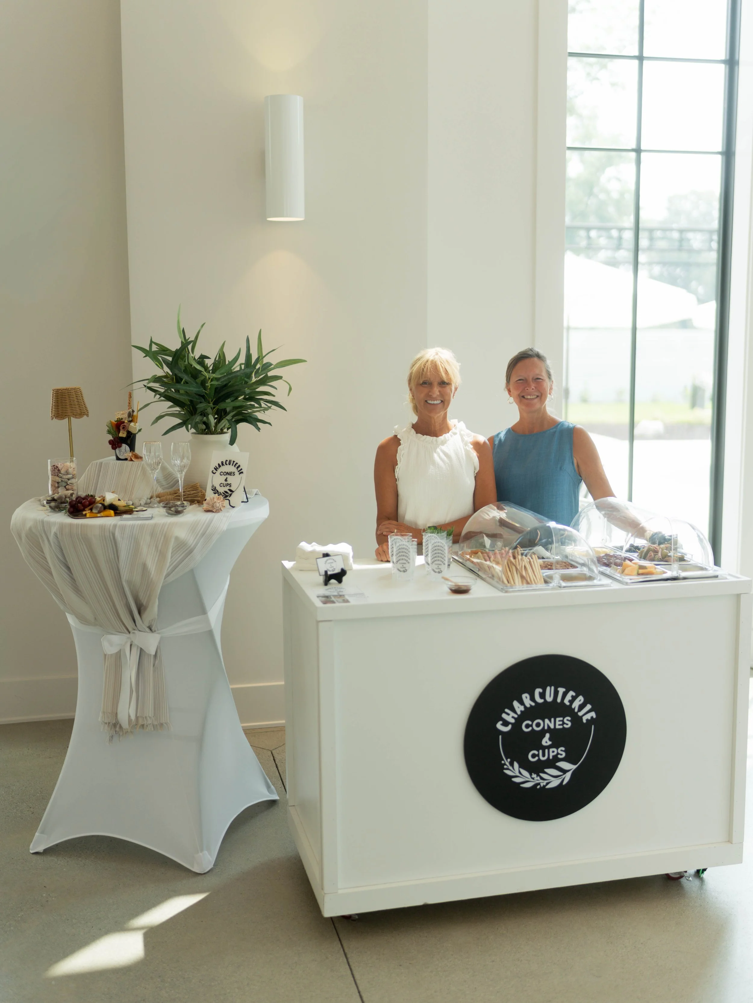 Two smiling women standing behind a white promotional table with a black circular sign that reads 'CHARCUTERIE CONES & CUPS' at an indoor event with large windows and natural light.