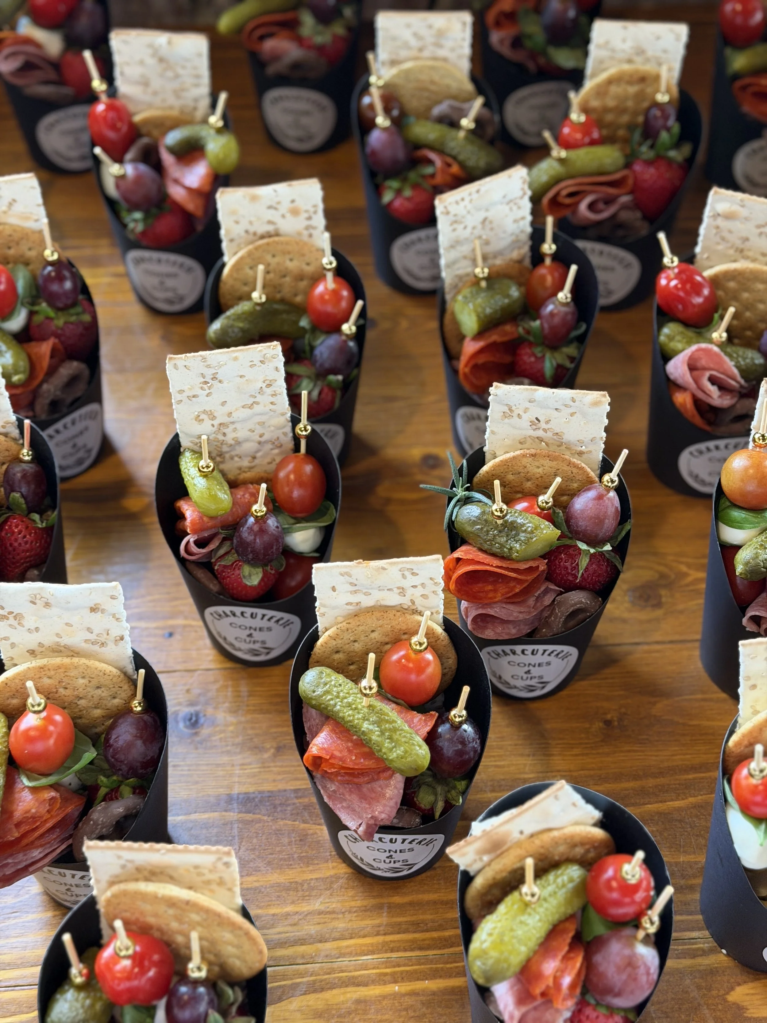 Multiple black cups filled with assortment of snacks including cherry tomatoes, slices of meat, pickles, crackers, strawberries, and cheese, placed on a wooden table.