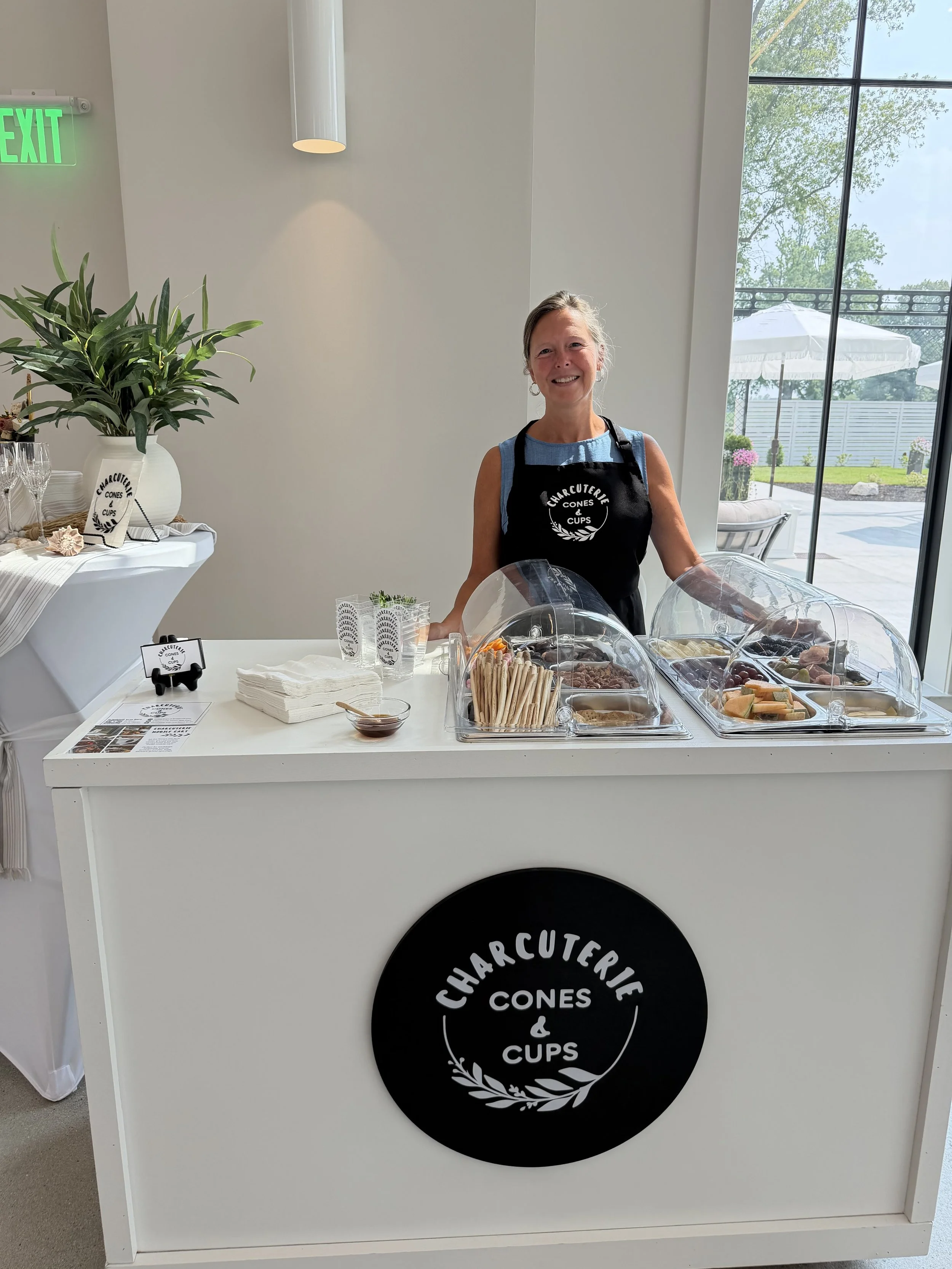Smiling woman in apron standing behind a white booth selling charcuterie cones and cups, with a large window showing an outdoor patio with umbrellas in the background.