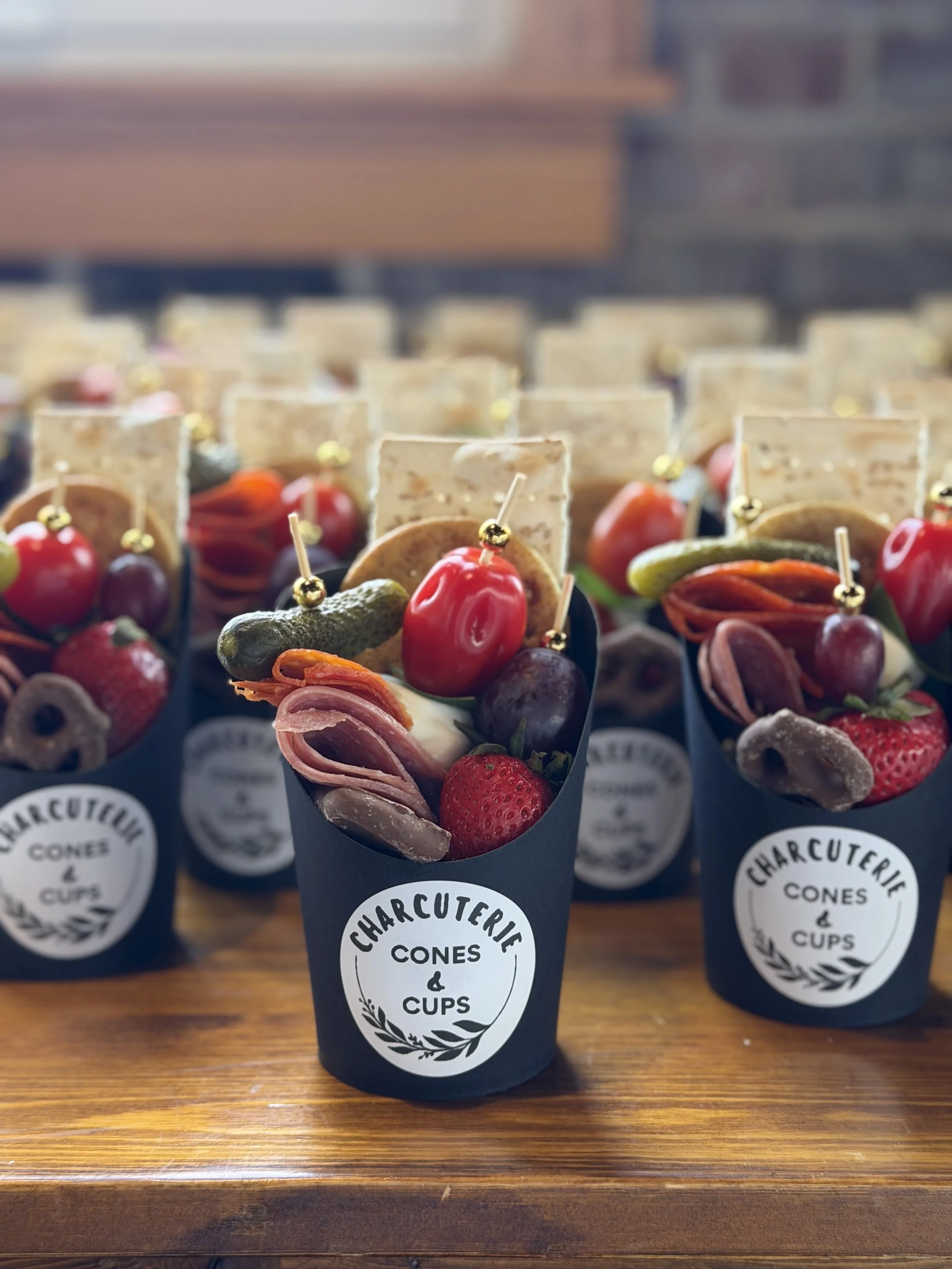 Assorted charcuterie cones with meats, cheeses, grapes, strawberries, pickles, and crackers wrapped in black paper cups labeled "Charcuterie Cones & Cups" on a wooden surface.