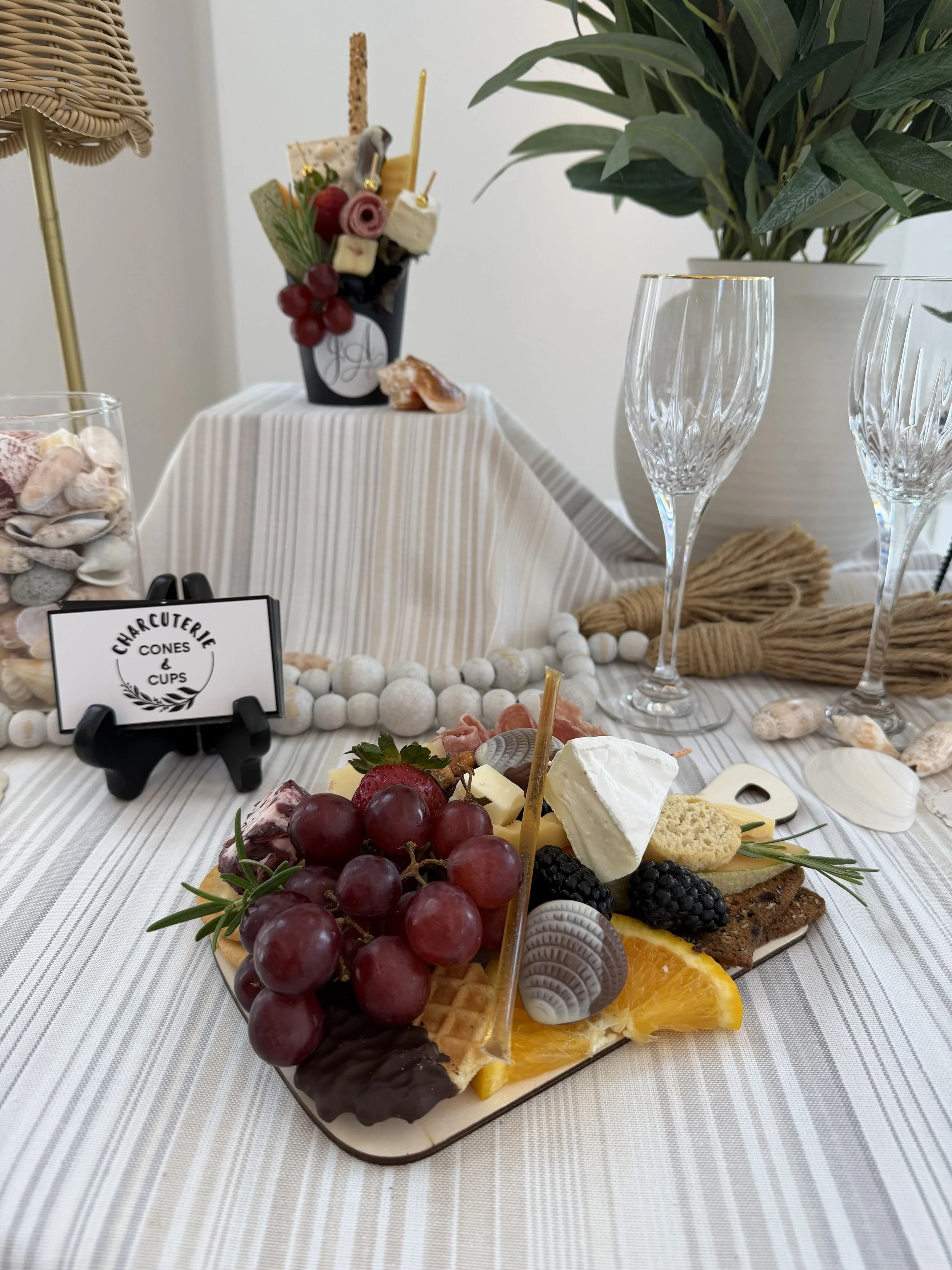 A dessert platter with grapes, blackberries, cheese, sliced oranges, cookies, and chocolate decorations on a white striped tablecloth, with a background of glass champagne flutes, a potted plant, and a sign reading 'Charcuterie Cones & Cups'.