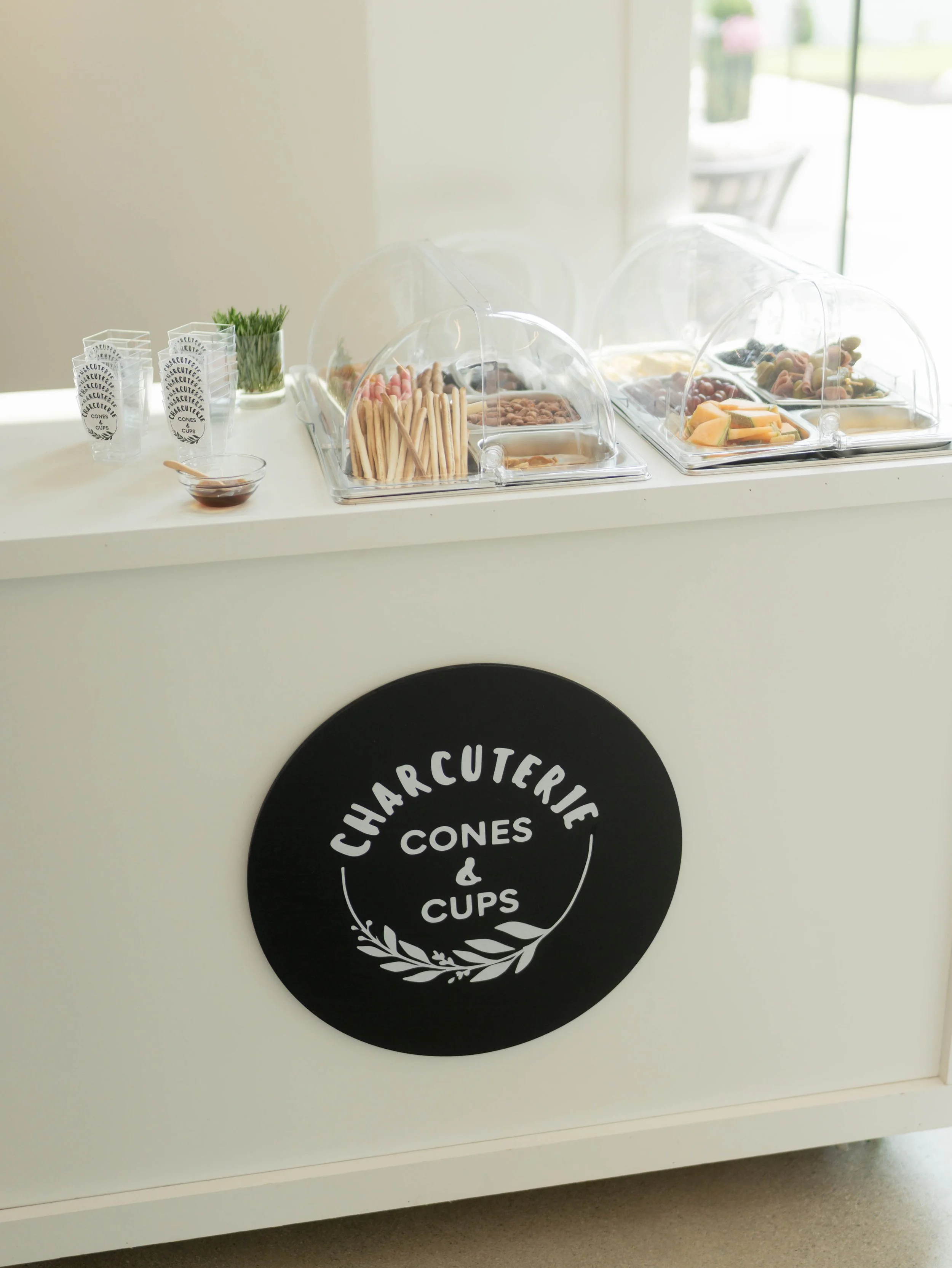 A white counter with clear plastic containers filled with snacks, a small bowl of syrup with a spoon, a glass of herbs, and a group of paper cups with a logo that reads 'Charcuterie, Cones & Cups' in black circle.